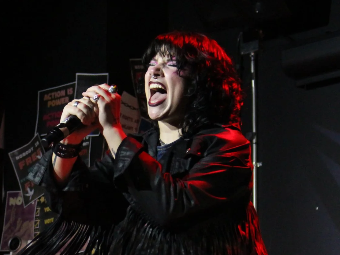 A woman with black hair, glasses, and fishnet stockings performing passionately on stage, holding a microphone with her mouth open.