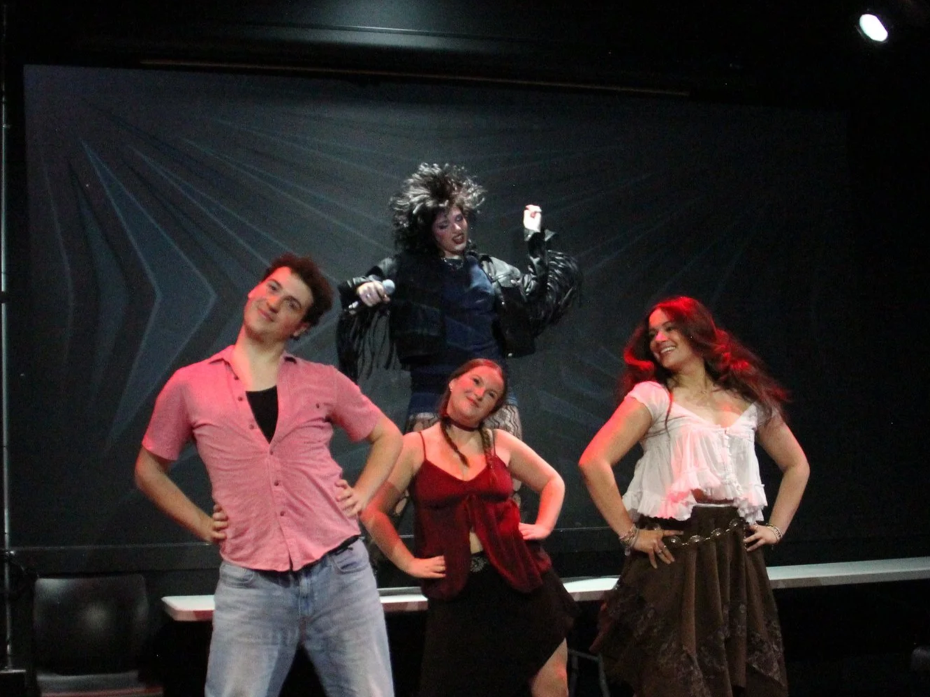 Four young performers on stage, three women and one man, striking confident poses with the woman at the back holding a microphone. The background is black with stage lighting.