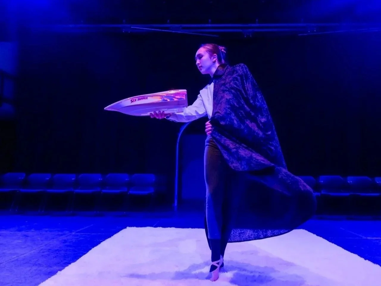 Woman holding a toy boat on a stage with blue lighting, standing on a white rug.