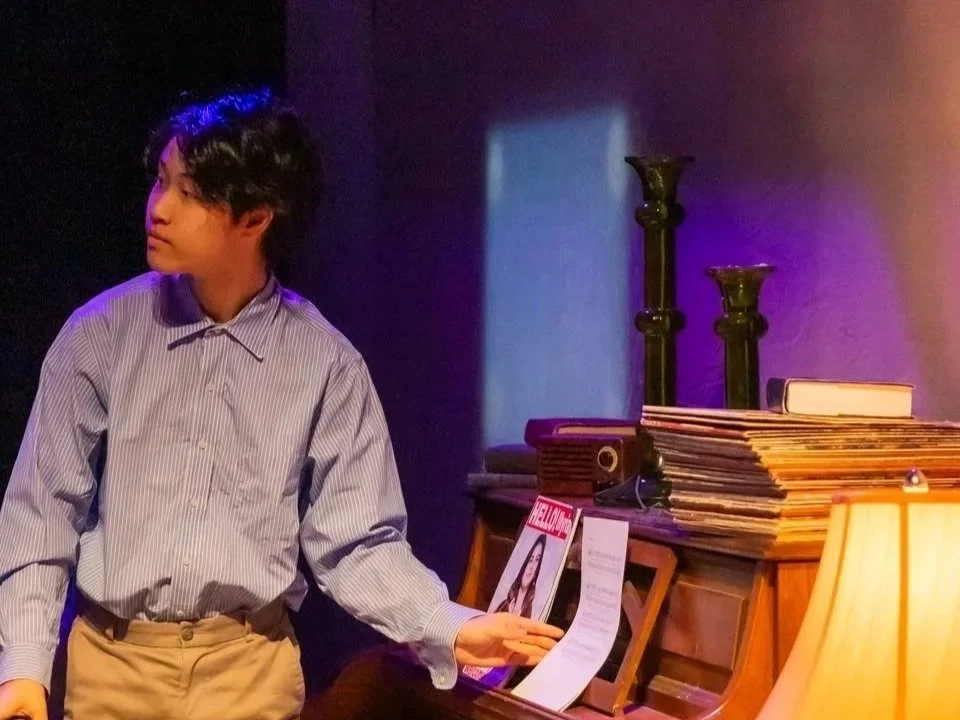 A young man in a striped shirt and khaki pants standing next to a wooden piano, looking to the side, with magazines and a record on top, in a dimly lit room with a table lamp.