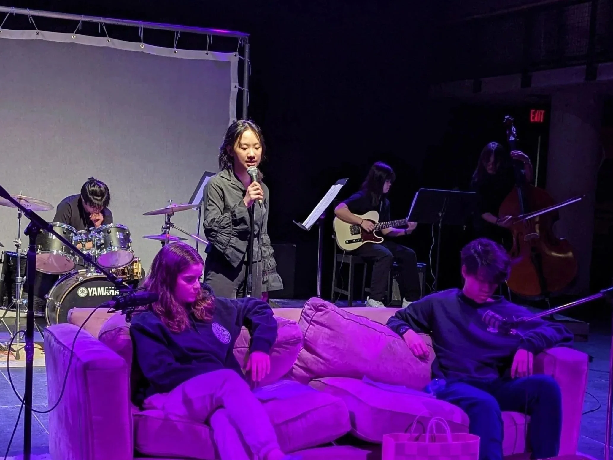 A group of young musicians performing on stage, with a woman singing into a microphone in front of an audience. The band includes a drummer, a guitarist, and a double bassist, with the performers behind the singer. The stage is dimly lit with purple 