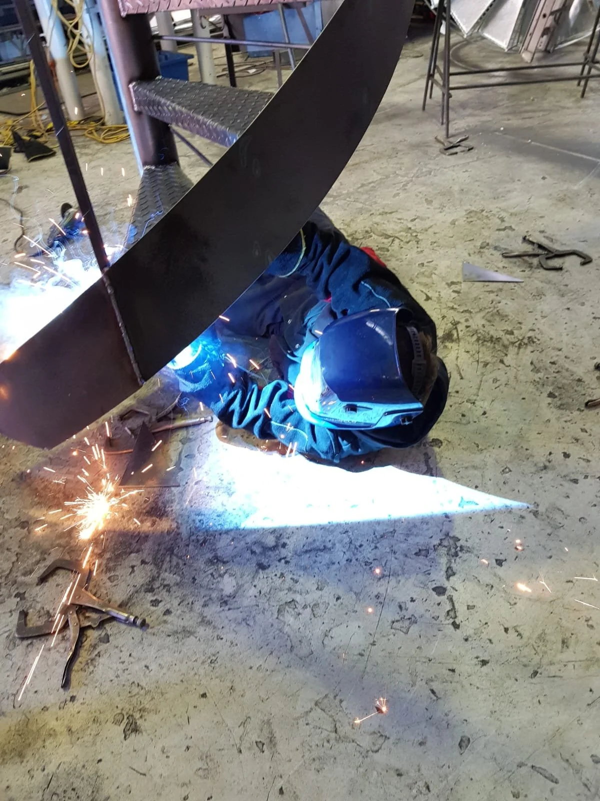 A person welding a large black metal piece while lying on the floor, wearing a welding helmet and protective clothing. Sparks are flying around in a workshop or industrial setting.