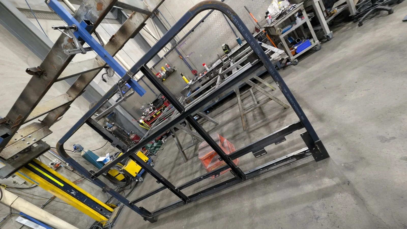 Metal framework and tools in a workshop or factory setting with workbenches, shelves, and equipment.