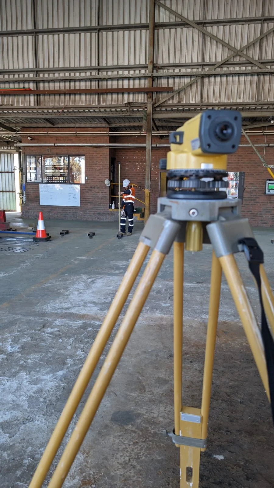 Construction site with a yellow tripod-mounted surveying instrument in the foreground, a construction worker in safety gear in the background, and a brick building with a window, wall-mounted notice, and cones on the ground.
