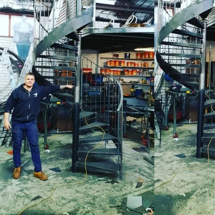 A person standing next to a metal spiral staircase inside an industrial or workshop setting with tools and supplies visible in the background.