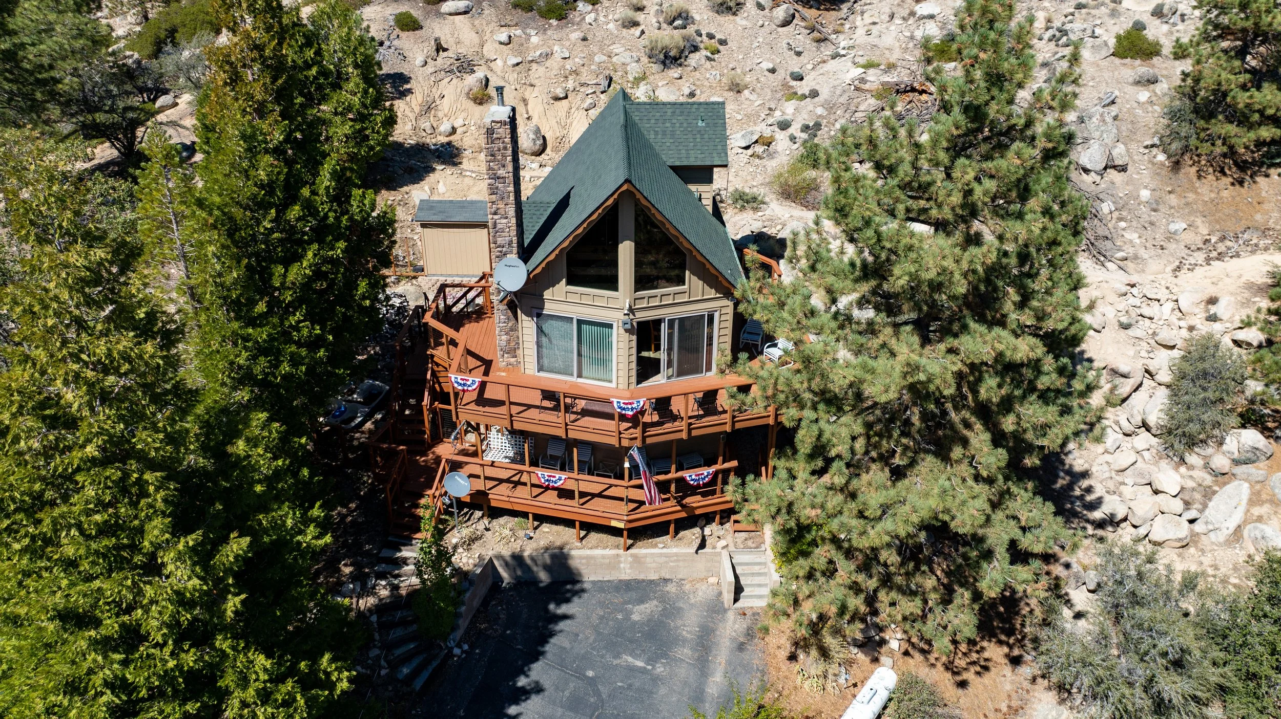 Aerial view of a house surrounded by tall trees with a large deck decorated with red, white, and blue bunting and an American flag, set in a rocky, natural landscape.