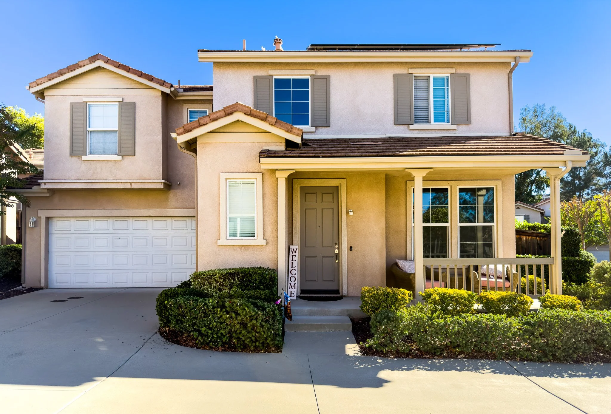 Front view of a two-story beige house with a garage, porch, and well-maintained bushes, under a clear blue sky.