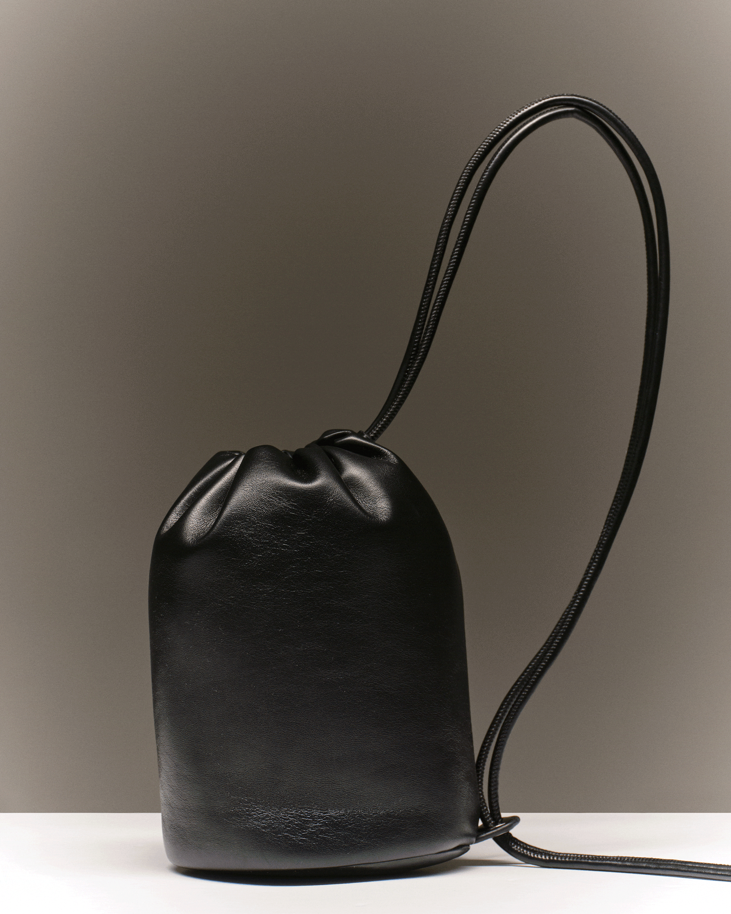 Black leather handbag with a long strap on a white surface against a gray background.