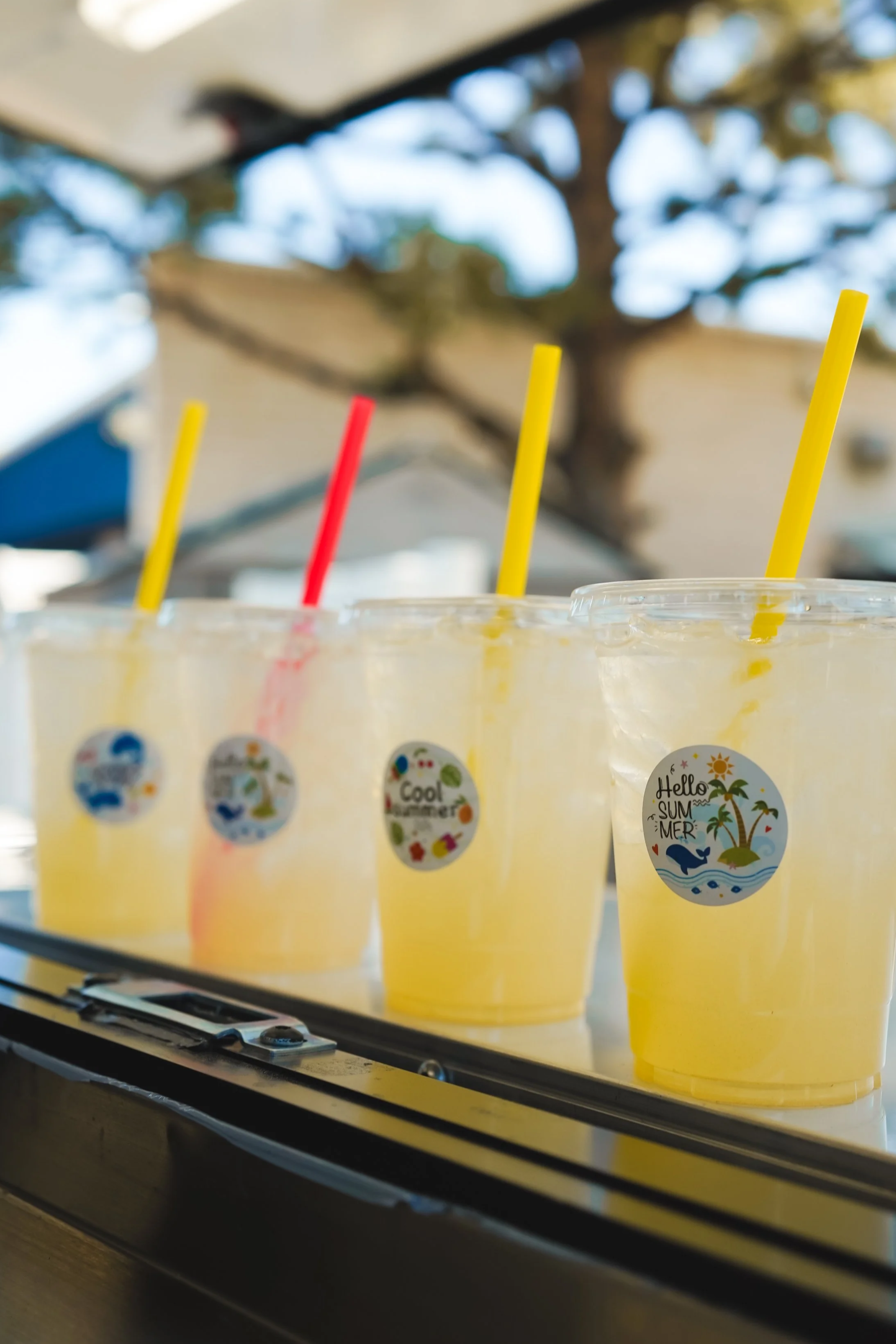 Two clear plastic cups with lemon slices, decorated with green and yellow star-shaped confetti, black straws, and green tinsel, on a table with a Buzz Lightyear-themed birthday party sign that reads 'Buzz's Blaster Punch Mango + Lilikoi Lemonade'.