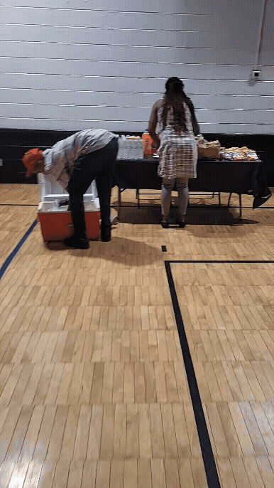 Two people are at a table with snacks and drinks in a gymnasium, one person is bent over a cooler, the other is standing and facing the table.