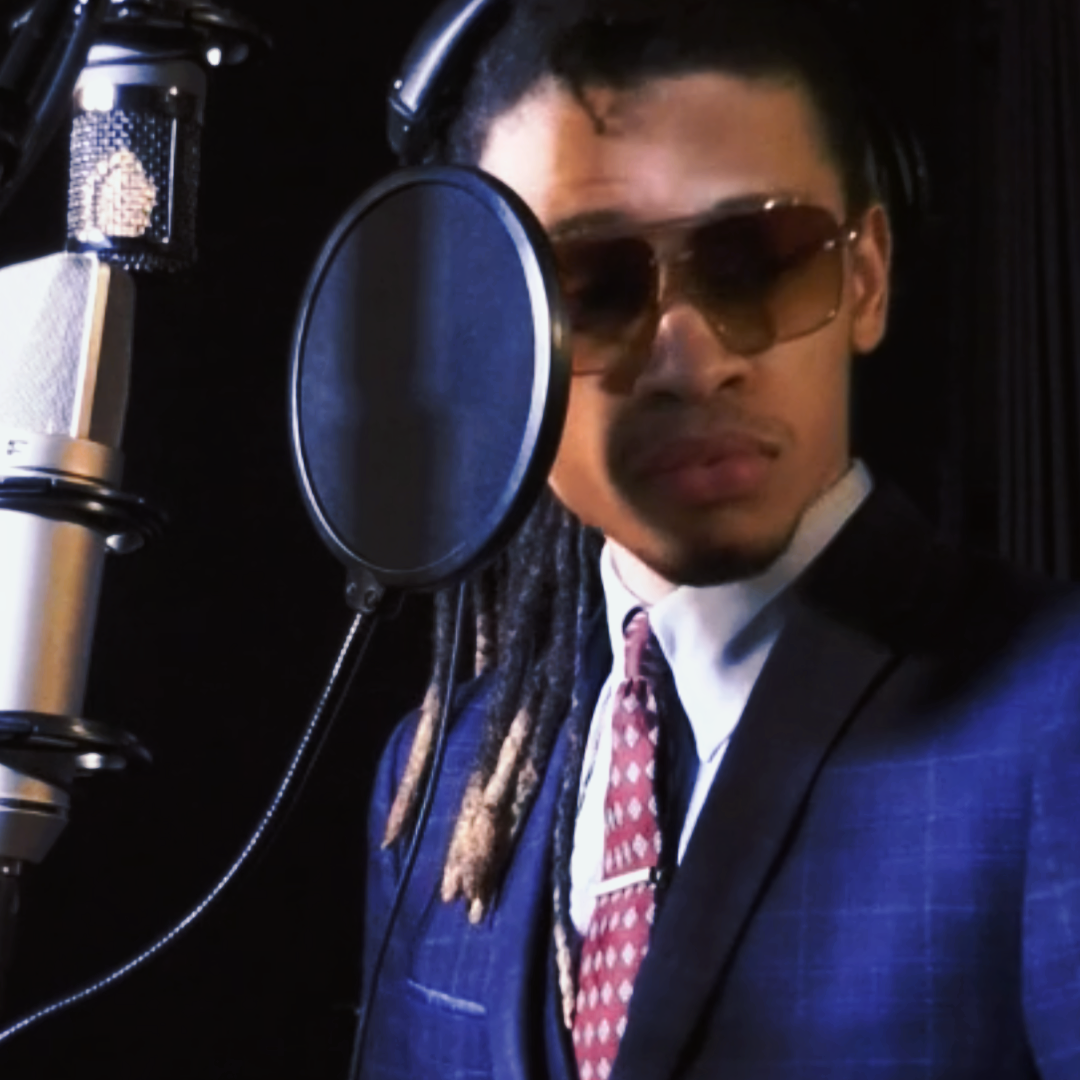 A man wearing sunglasses and a suit is in a recording studio, standing in front of a microphone with a pop filter.