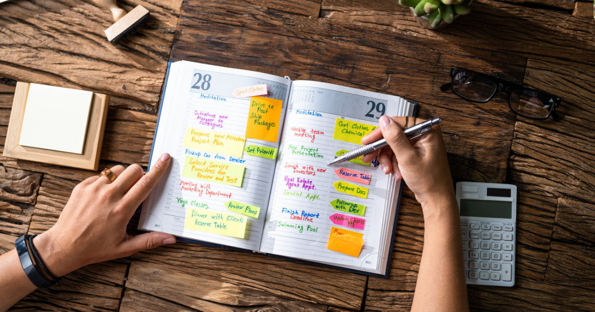 Hands writing in an organized daily planner with colorful sticky notes and a calculator, symbolizing business organization.