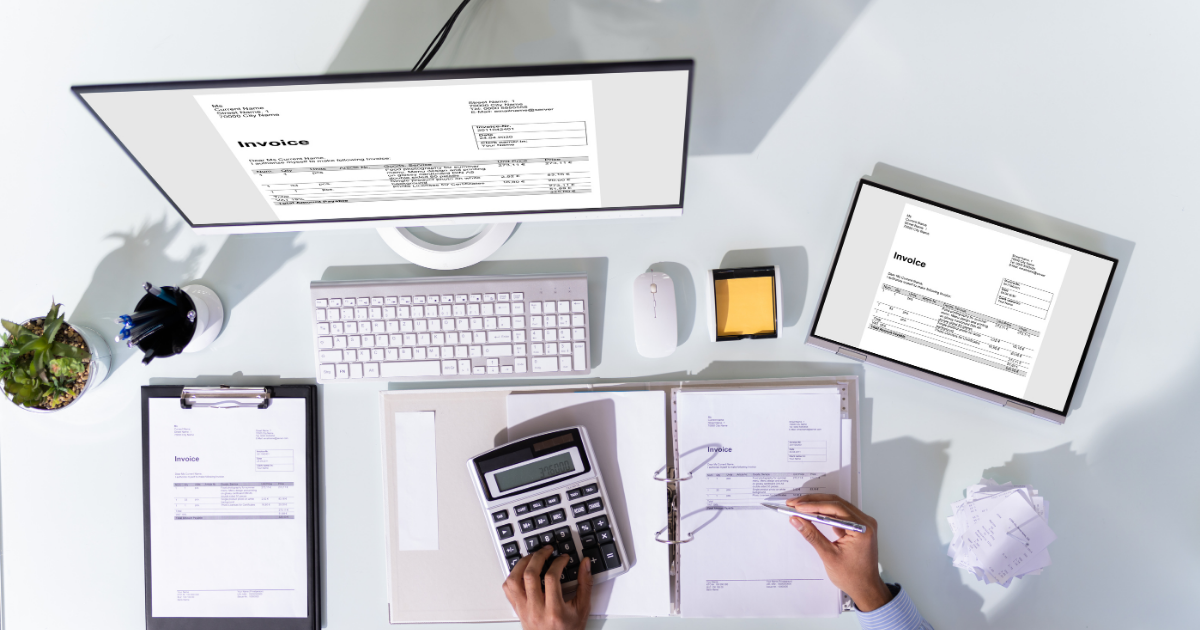 Overhead view of a desk with invoices, a calculator, a computer monitor, and a tablet showing business expense documents.