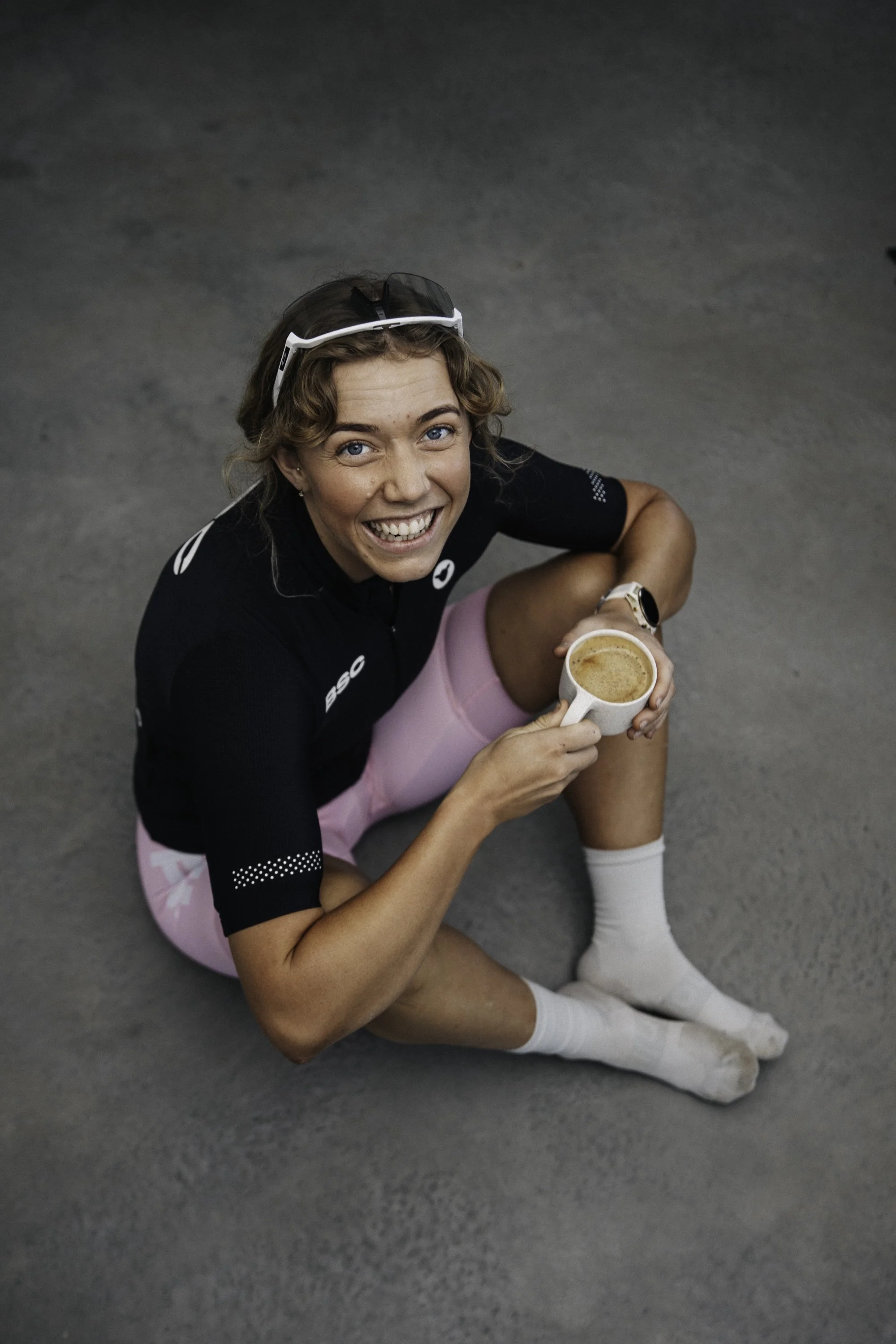 A smiling woman in athletic attire sitting on the ground, holding a cup of coffee, with sports sunglasses on her head and a smartwatch on her wrist.