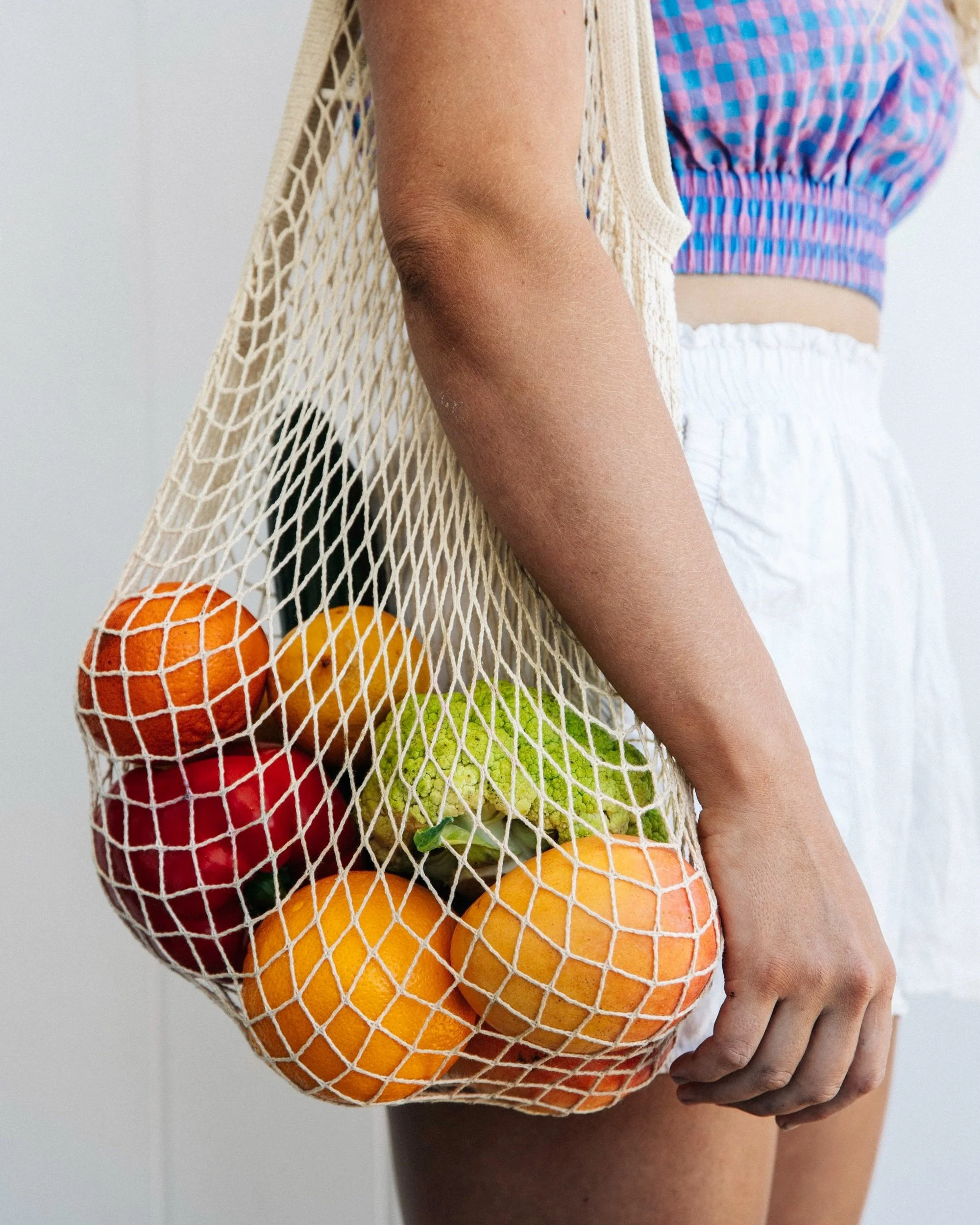 A person holding a mesh shopping bag filled with oranges, apples, and other fruits, wearing a colorful checkered crop top and white shorts.