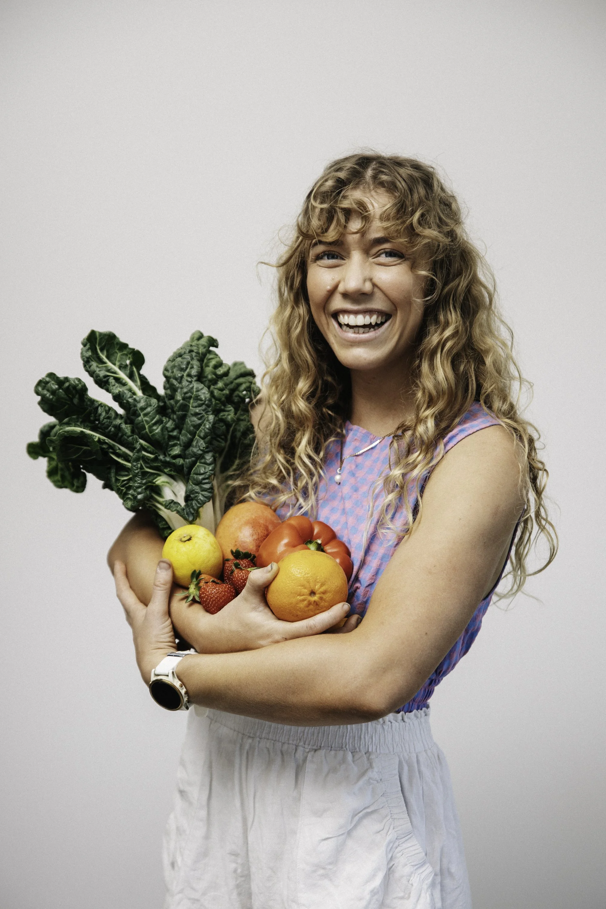 Woman smiling and holding fresh fruits and vegetables including leafy greens, strawberries, oranges, a lemon, a tomato, a peach, and an apple against a plain background.