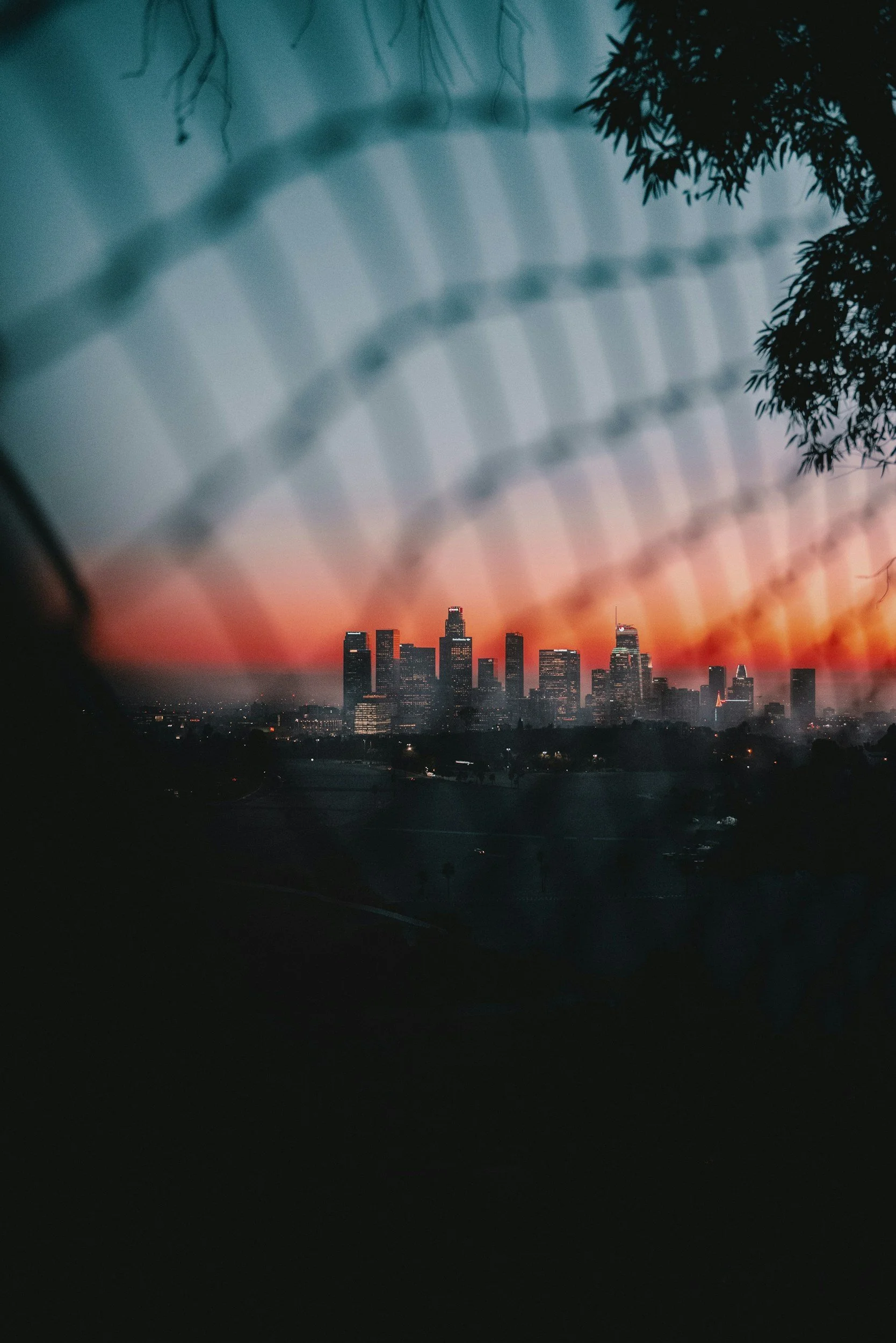 Silhouette of a city skyline at sunset, viewed through a chain-link fence, with the sky gradient from orange to blue, and silhouetted trees in the foreground.