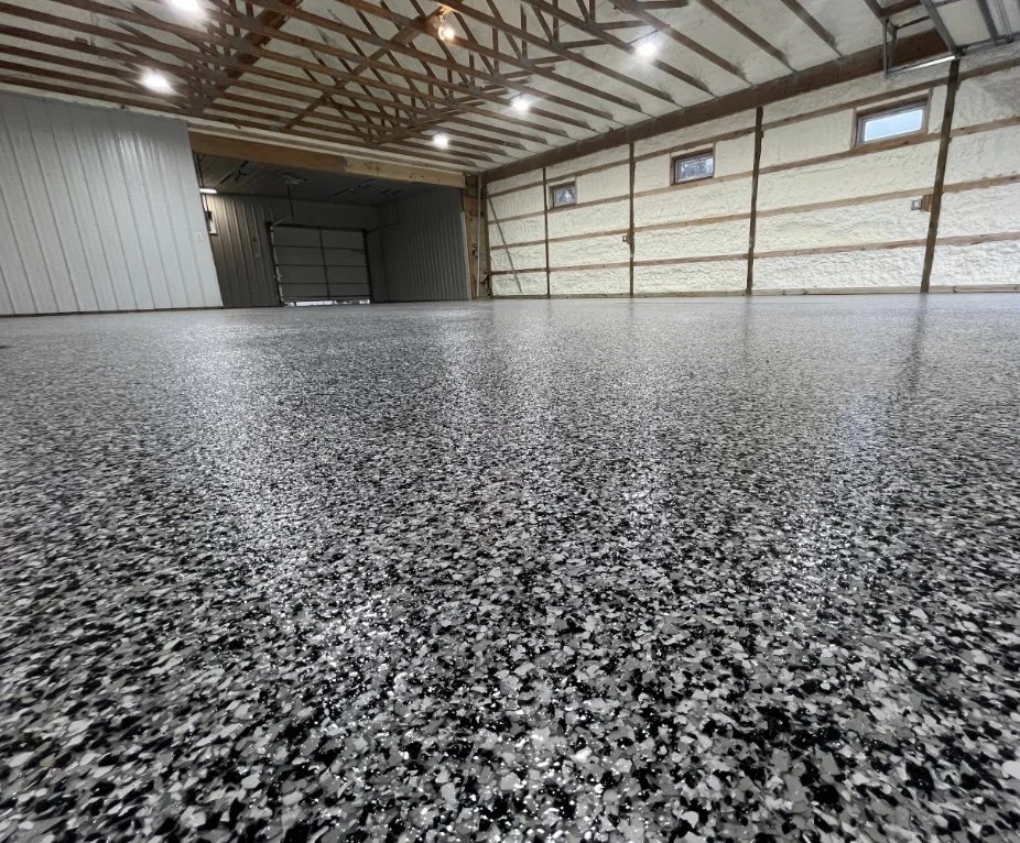 Empty garage with polished speckled gray epoxy floor, wooden ceiling beams, and unfinished insulated walls with small windows.