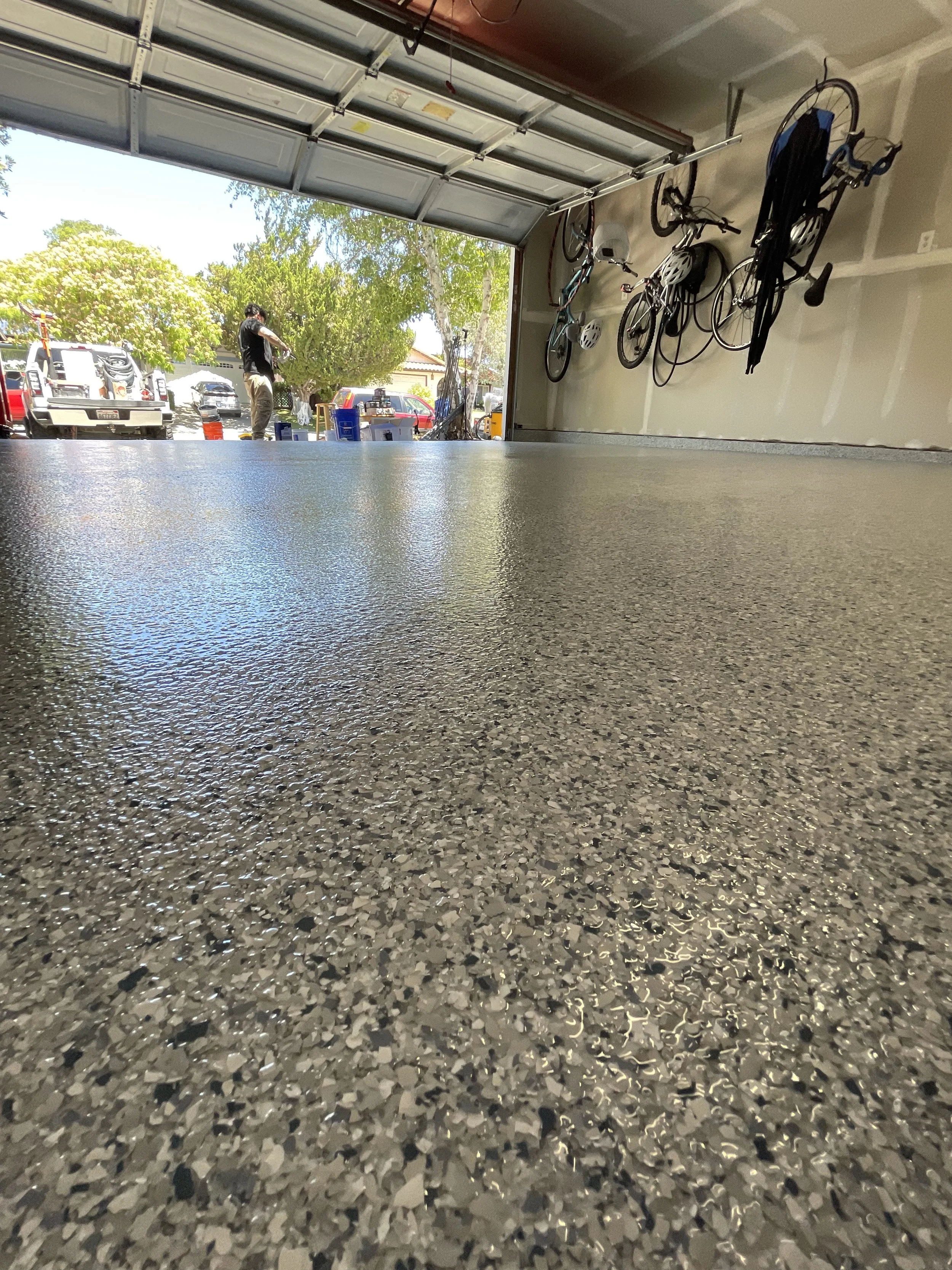 View from the floor inside a garage looking out through the open door. Bicycles hang from the wall on the right side. Outside, several vehicles are parked, and a person is standing near the trees and parked cars.