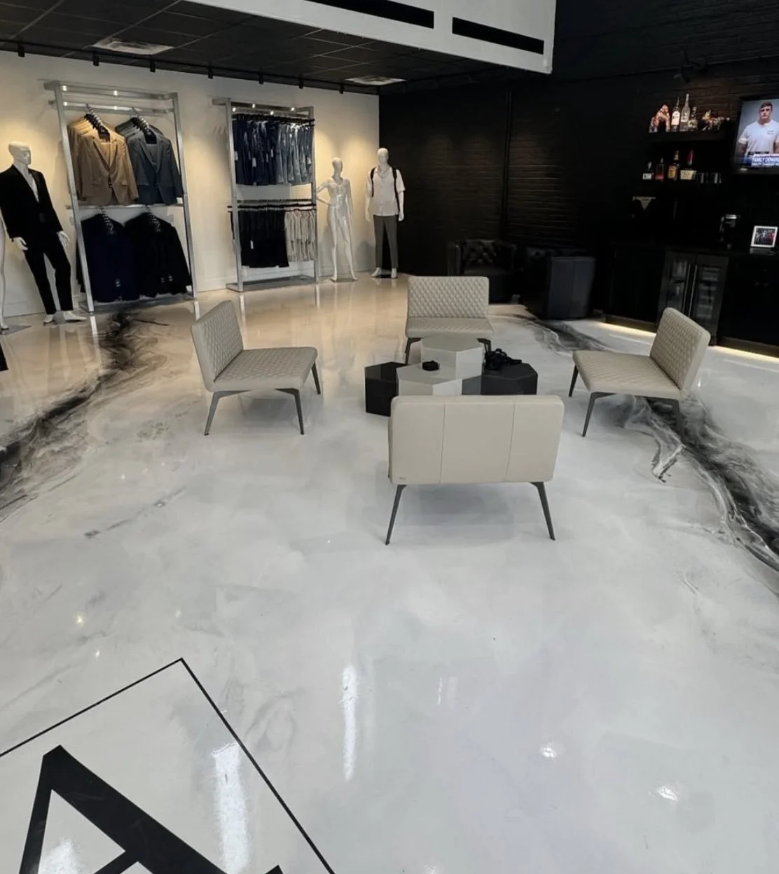 Inside a modern clothing store with mannequins displaying men's clothing, and a seating area with four beige chairs and black tables. The store features a marble floor with black and white veining, and a black wall with a TV and bar area.