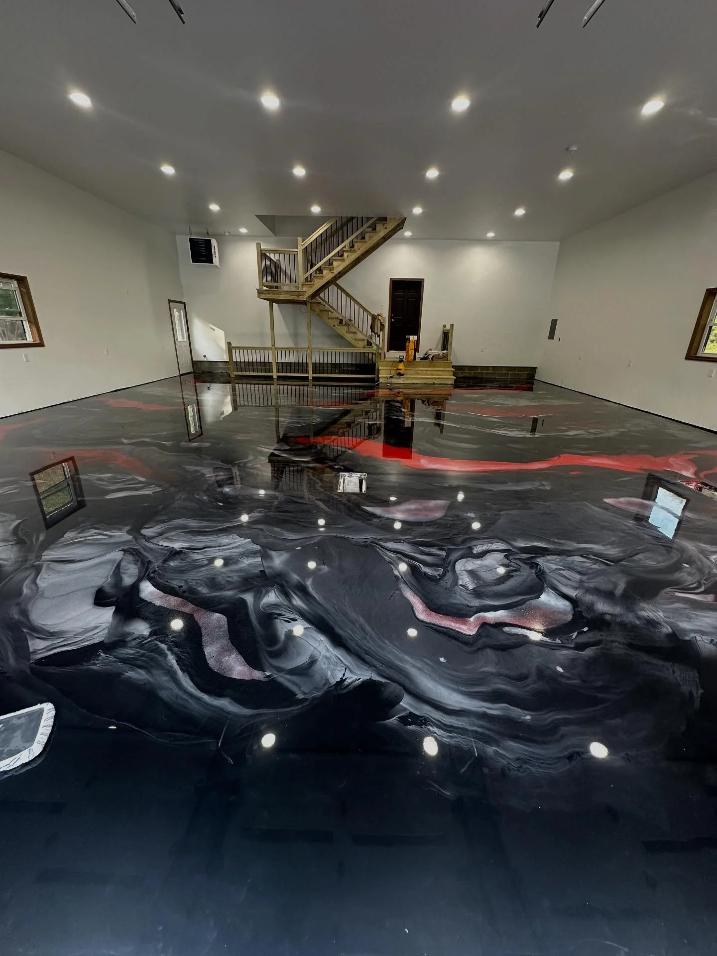 Empty room with glossy black and gray marbled flooring, white walls, multiple ceiling lights, and a wooden staircase leading to an upper level.