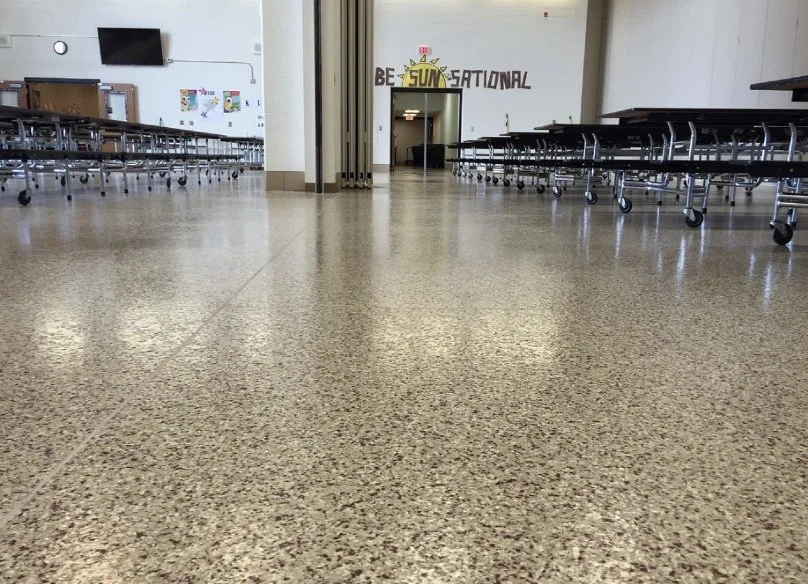 Empty school cafeteria with rows of stacked lunch tables on wheels, beige speckled flooring, a wall with colorful decorations, and a hallway entrance with the words 'BE SUN-SATIONAL' above it.