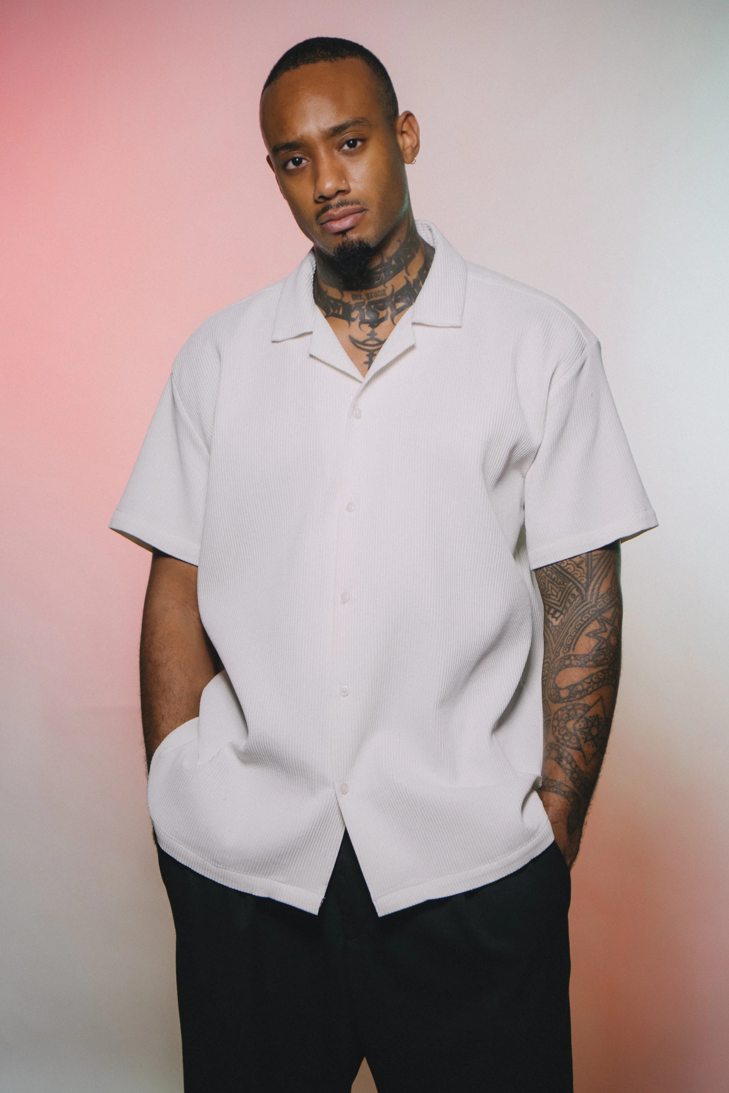 A young man with tattoos, wearing a white short-sleeved shirt and black pants, standing with hands in pockets against a pink and light gray background.