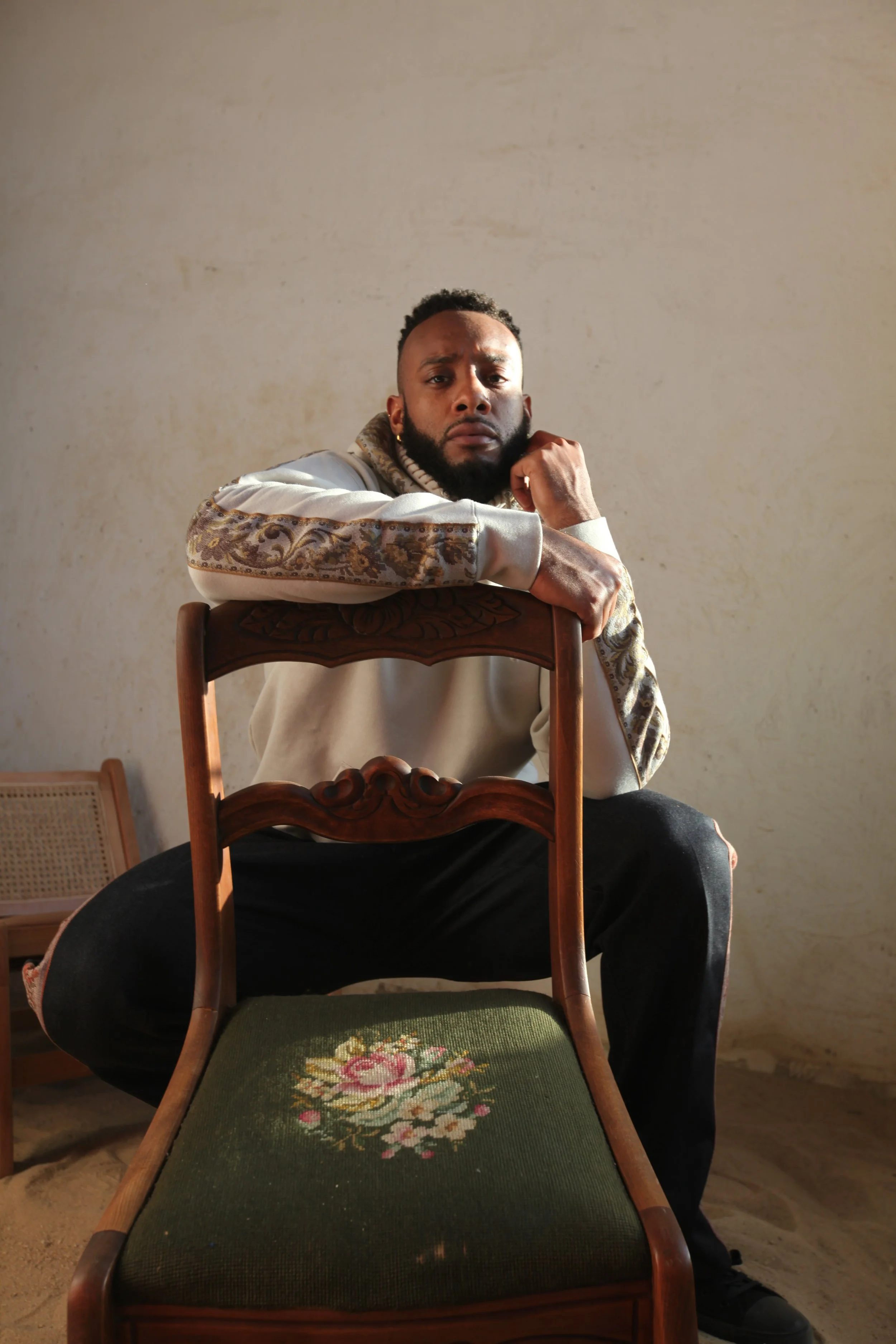 A man with a beard and short curly hair, wearing a beige sweatshirt with patterned sleeves, sits behind a wooden chair with floral upholstery, resting his arms on the chair's backrest, in a room with a plain beige wall.