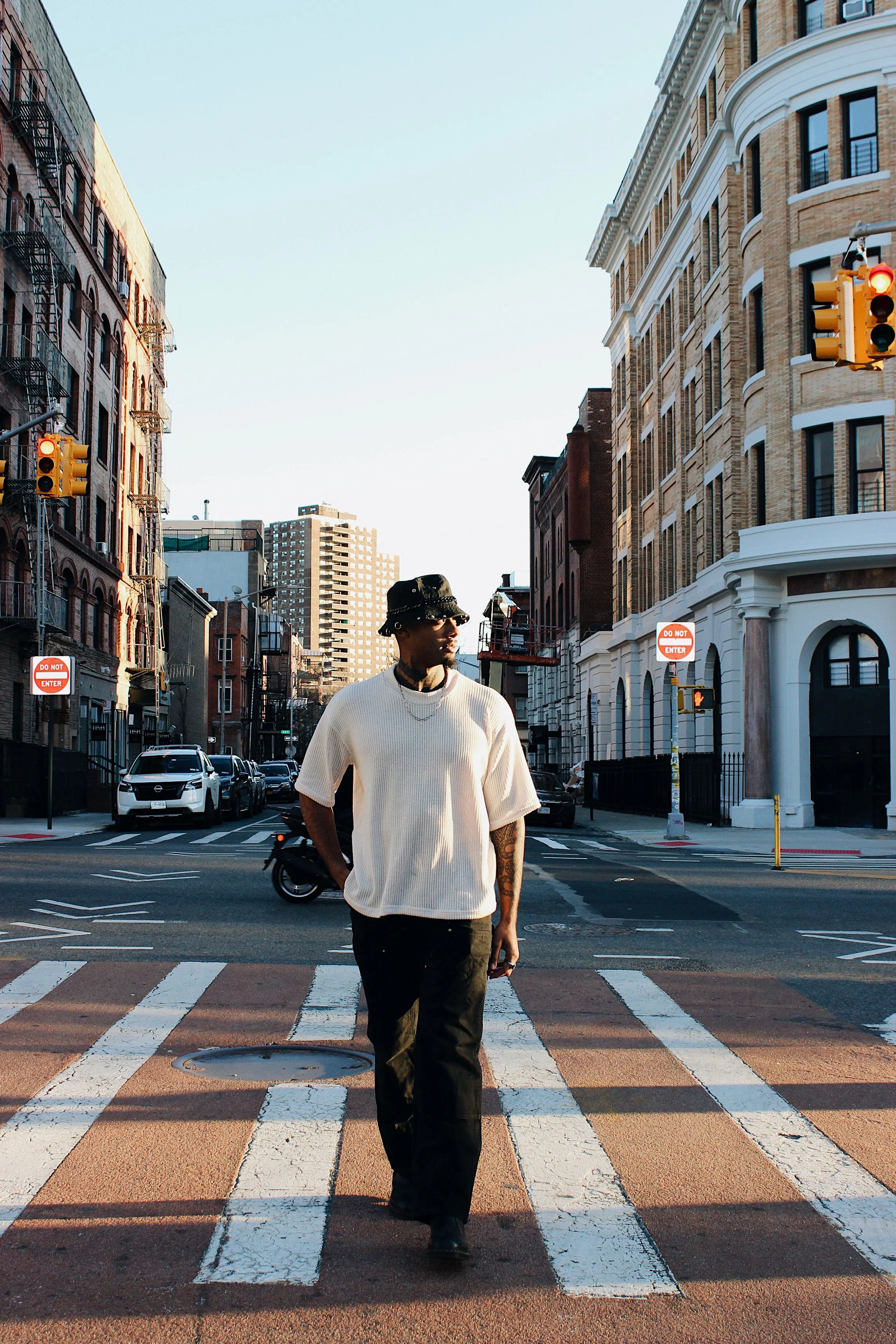 A man walking across a city street at crosswalk in the early evening. He is wearing a white oversized sweater, dark pants, a bucket hat, chain necklace, and sunglasses. The cityscape includes tall brick buildings, parked cars, traffic lights, and a clear sky.