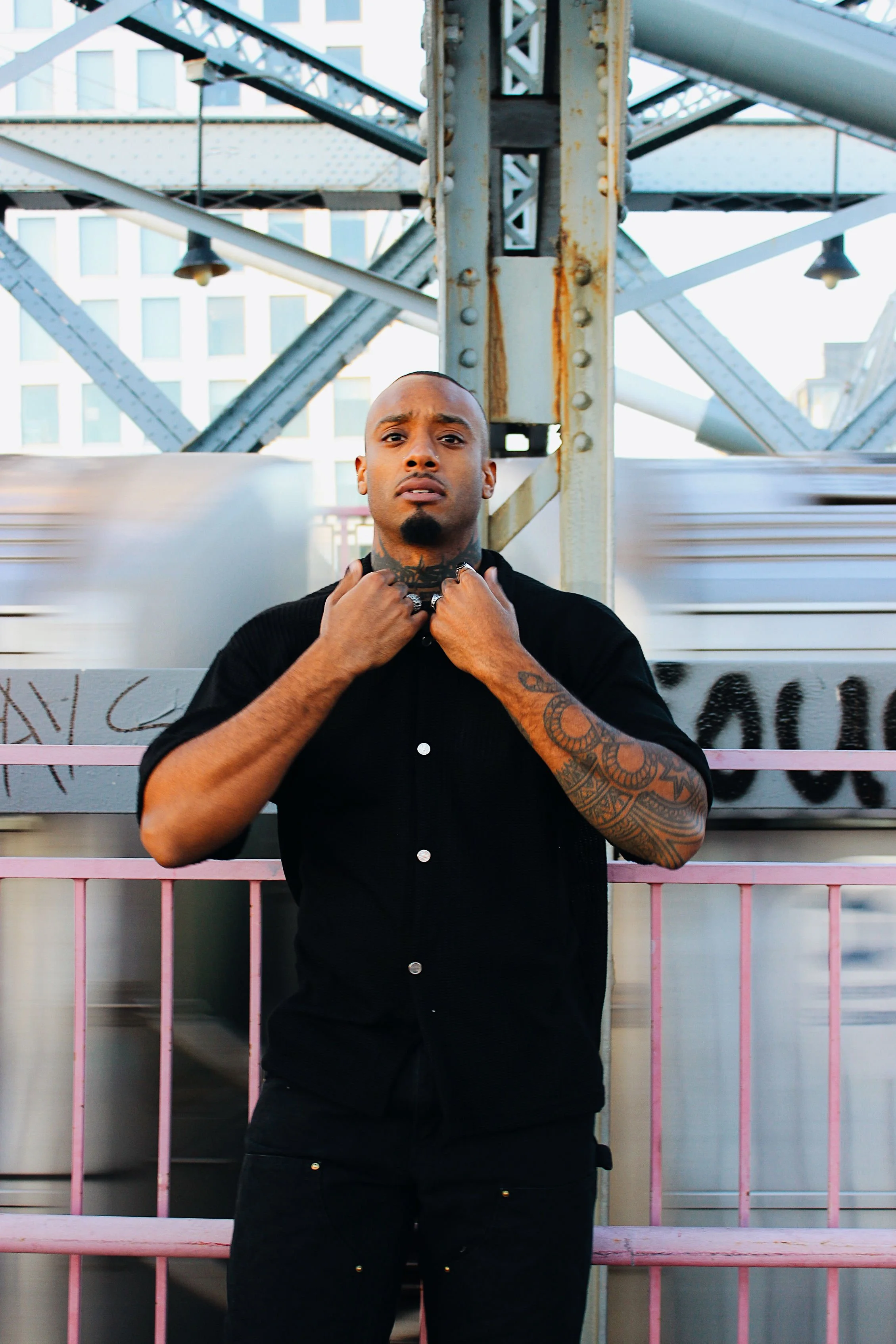 A man with tattoos on his arms and neck, wearing a black shirt, standing in front of a pink metal fence and a metal structure, possibly a bridge or tower, with blurred motion passing behind him.