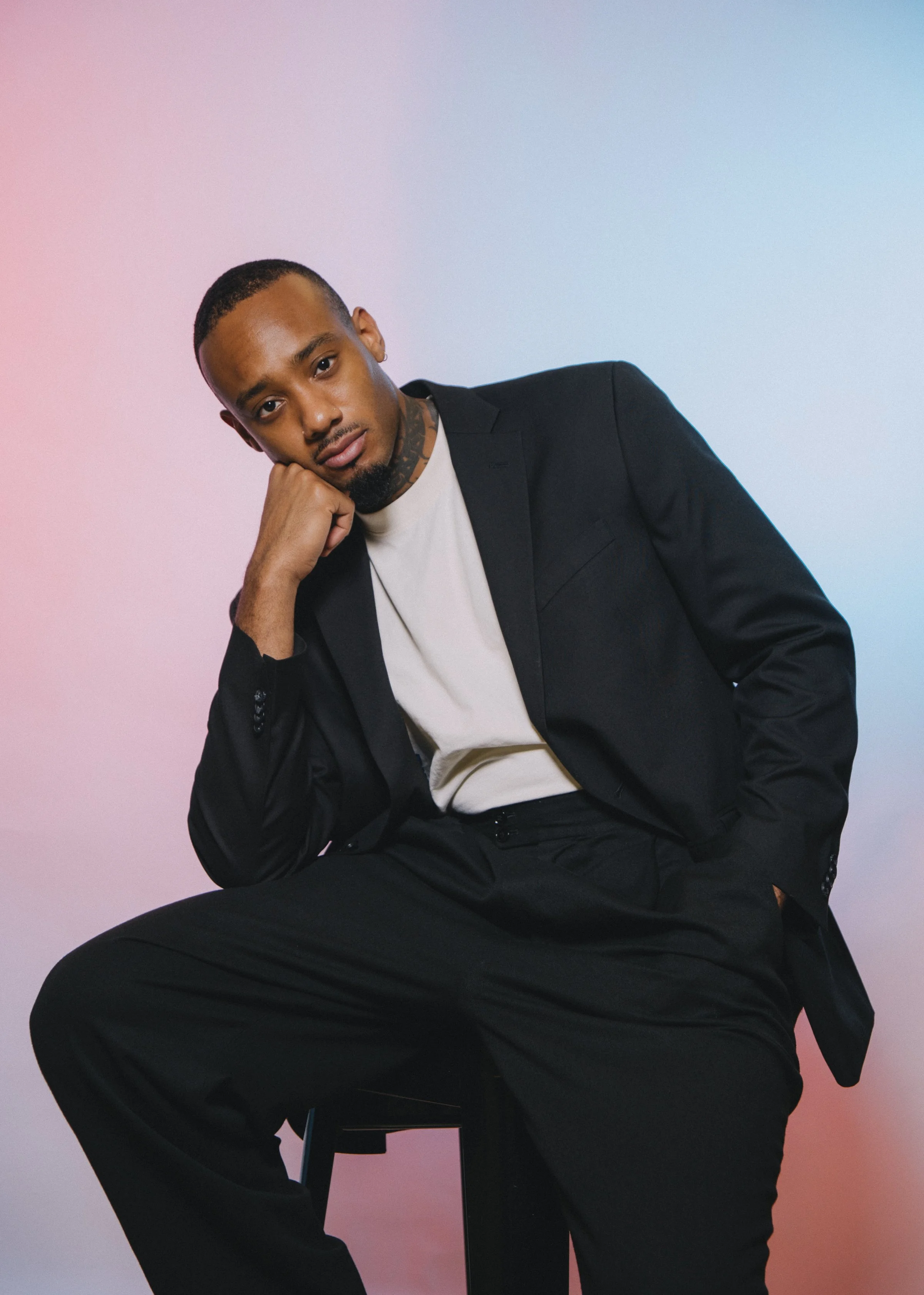 A man dressed in a black suit with a white t-shirt, sitting on a stool with his head resting on his hand, against a gradient pink and blue background.