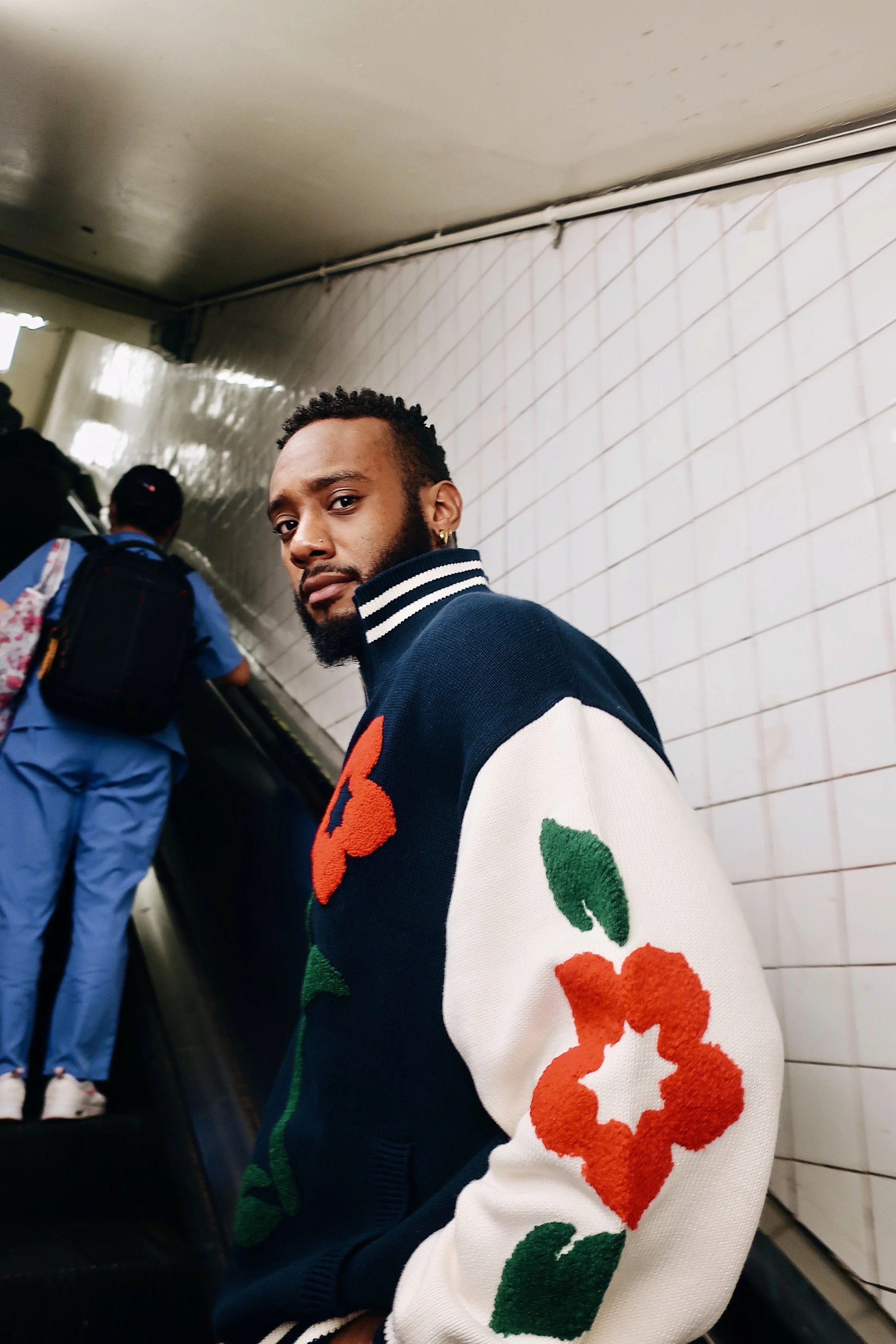 A young man with a beard and gold earrings looking back at the camera on an escalator in a subway station, wearing a navy and white varsity jacket with red and green floral details.