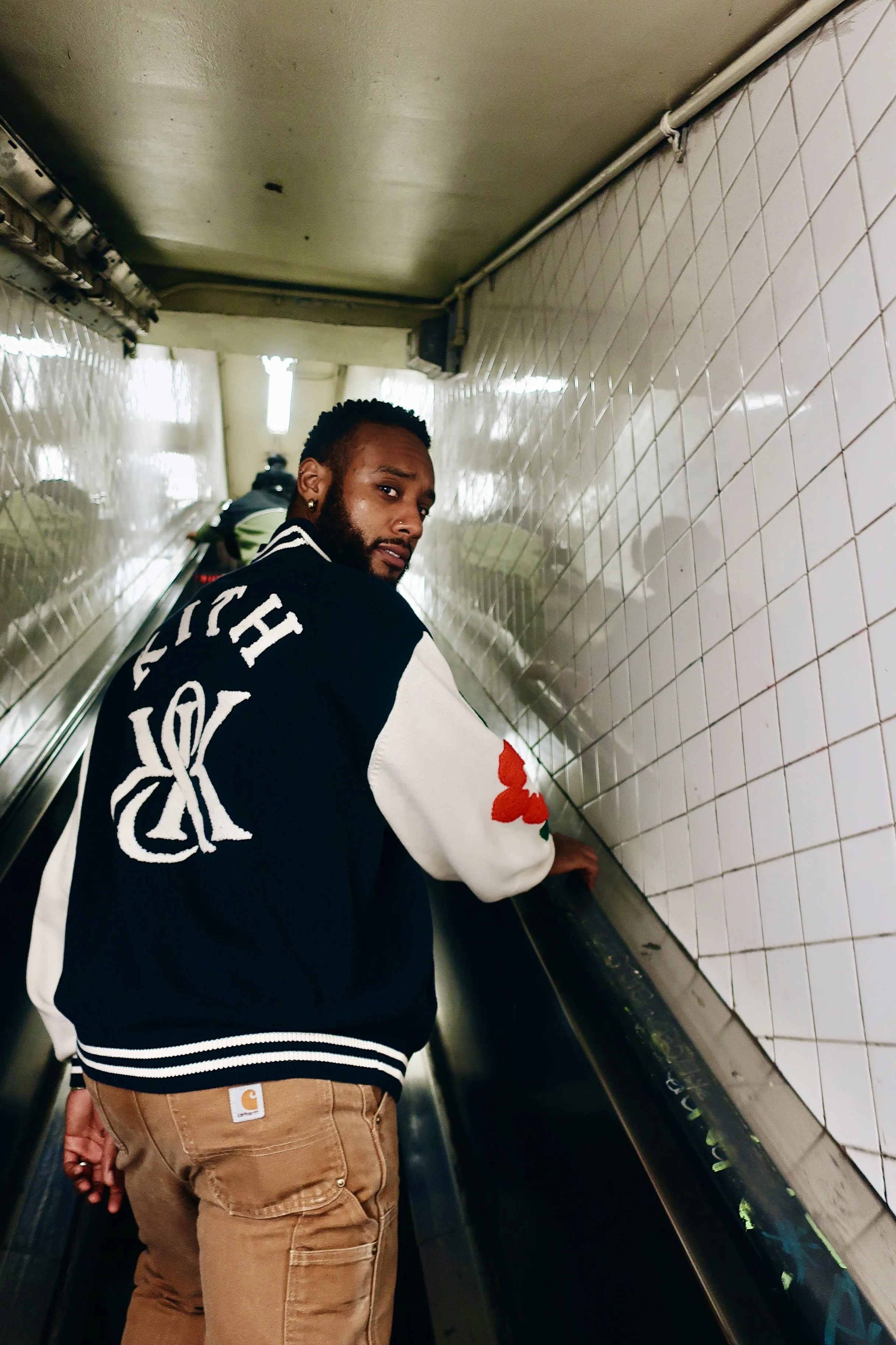 A man with a beard and earrings using an escalator in a subway station, wearing a black jacket with white sleeves and red floral patches