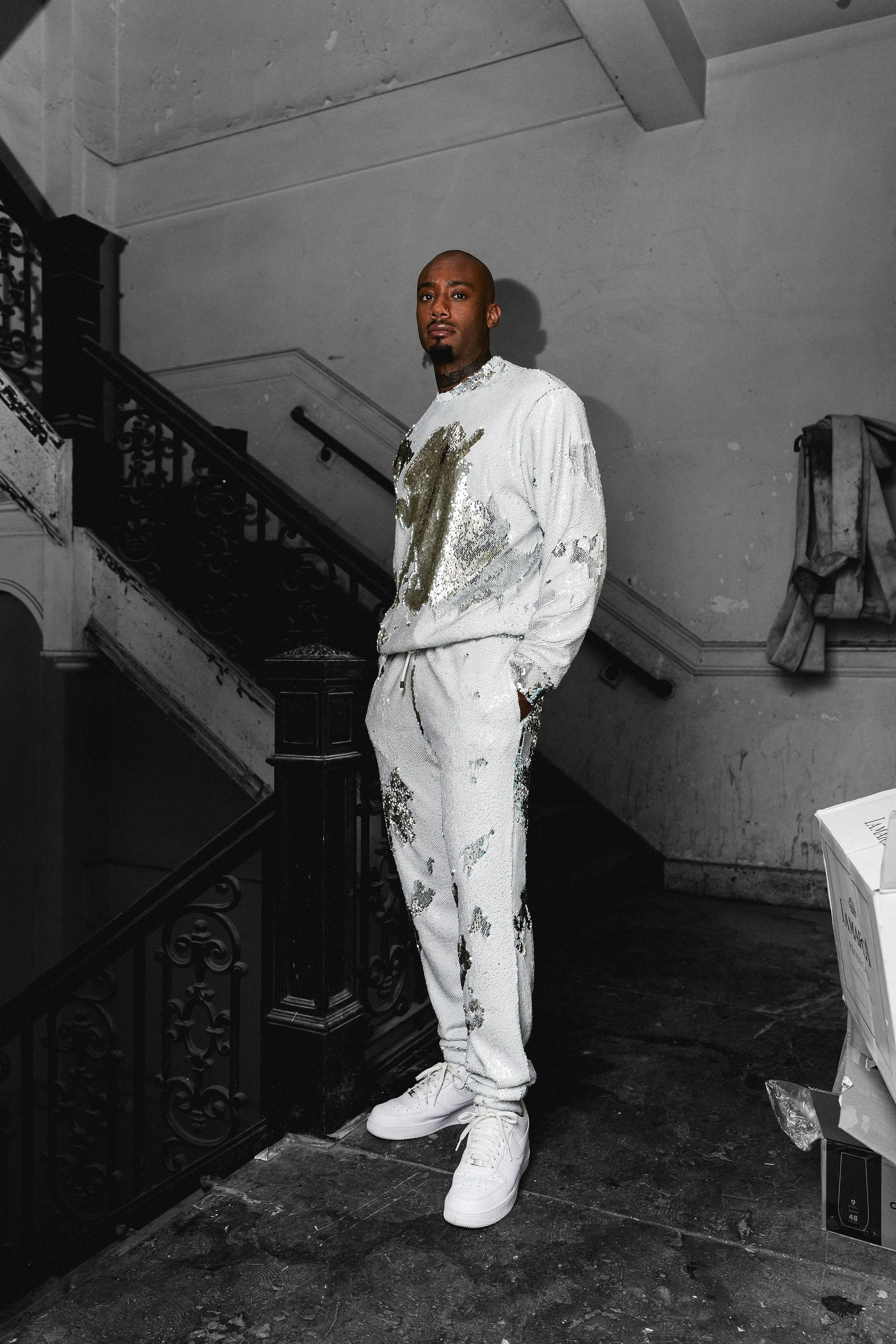 Man standing on a staircase landing in white clothing with gold and silver patterns, in an unfinished or abandoned building.