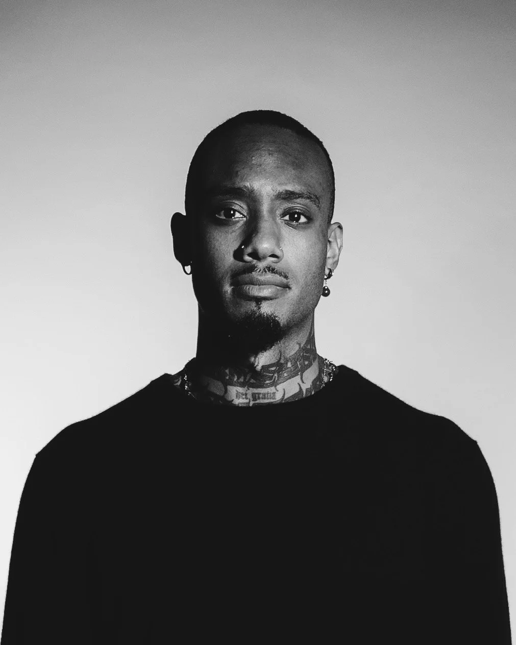 Black and white portrait of a young man with tattoos, earrings, and a nose ring, wearing a black shirt, against a plain background.