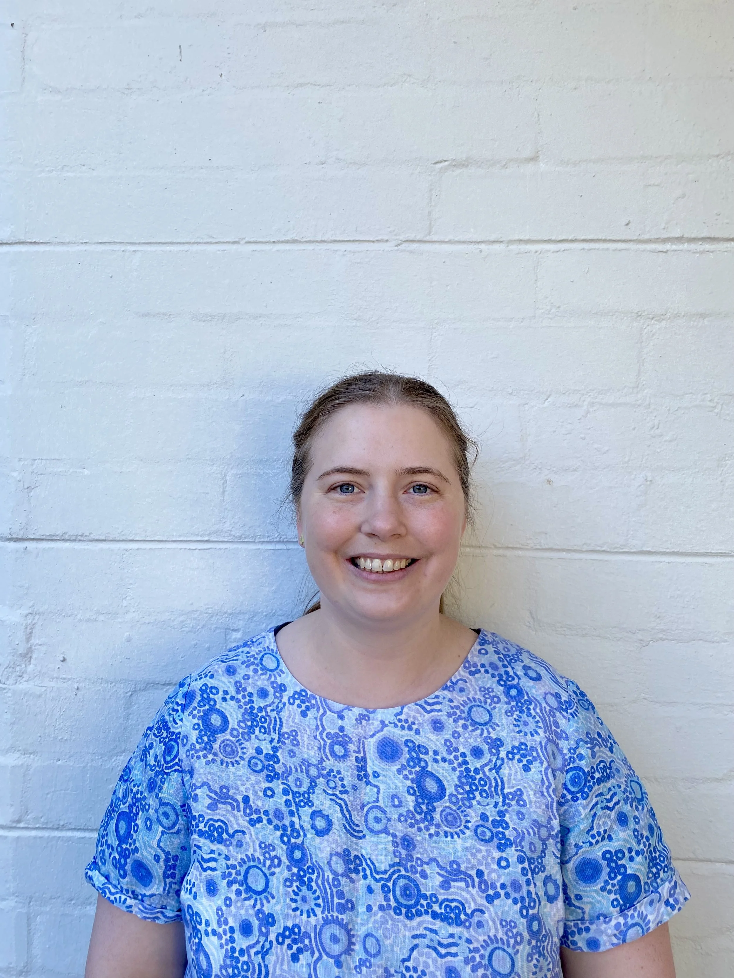 A smiling woman with light brown hair wearing a blue and white patterned top standing against a white brick wall.