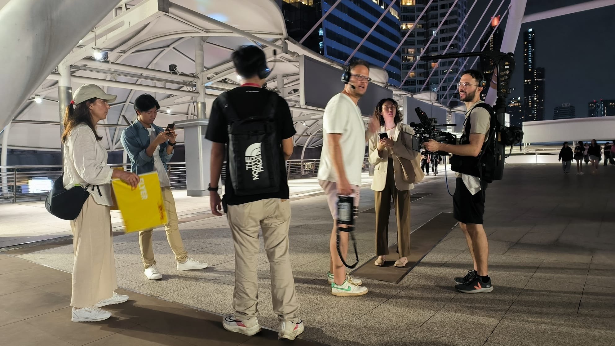 Film crew on urban walkway at night, using professional camera equipment and wearing casual clothing.