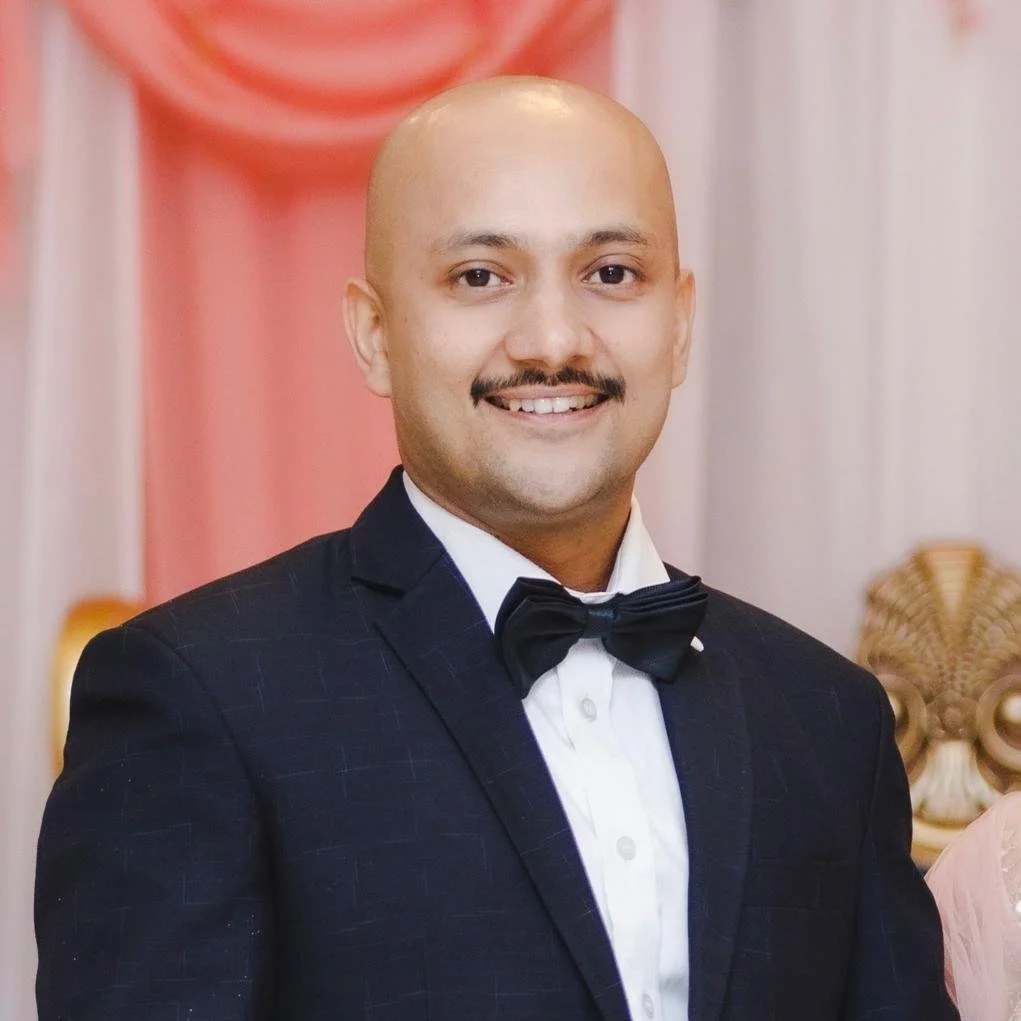 Man in a suit with a bow tie smiling in a formal setting with draped curtains in the background.