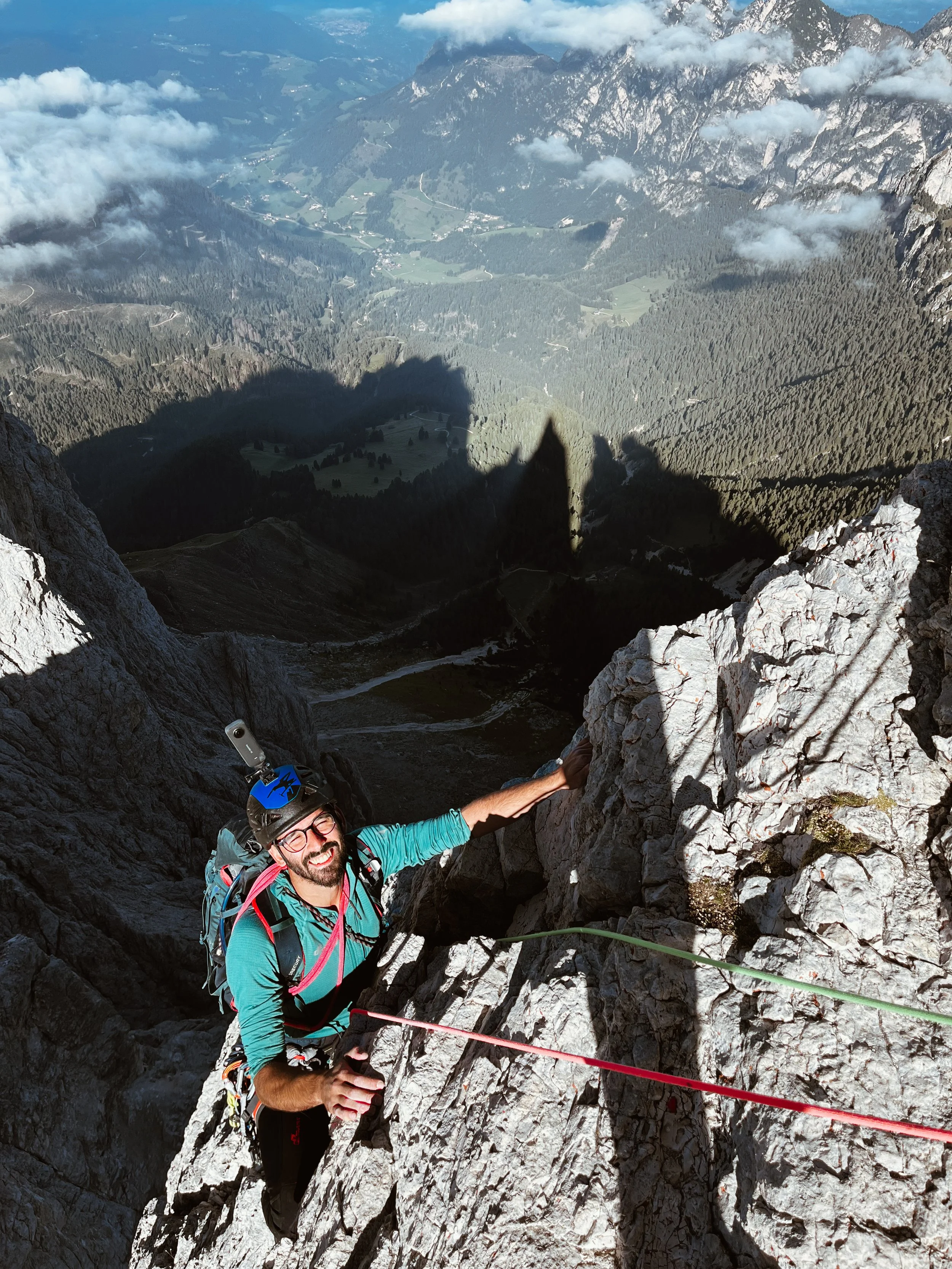 A man with a backpack and climbing gear, including a helmet, is climbing a steep rocky mountain, with a view of a valley and mountains below.