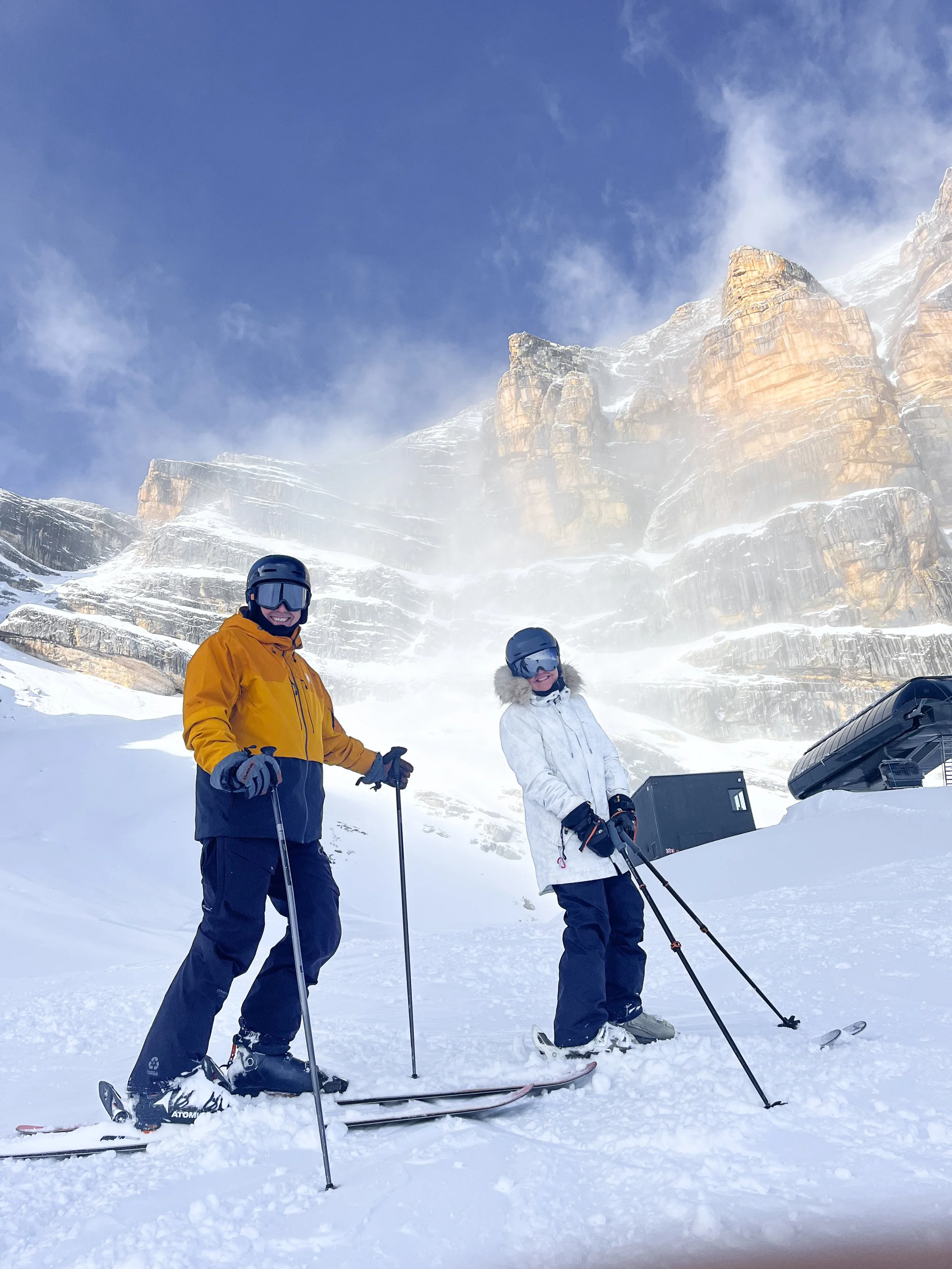 Two people dressed in winter gear and ski helmets standing on snow with cross-country skis and poles, mountains in the background.