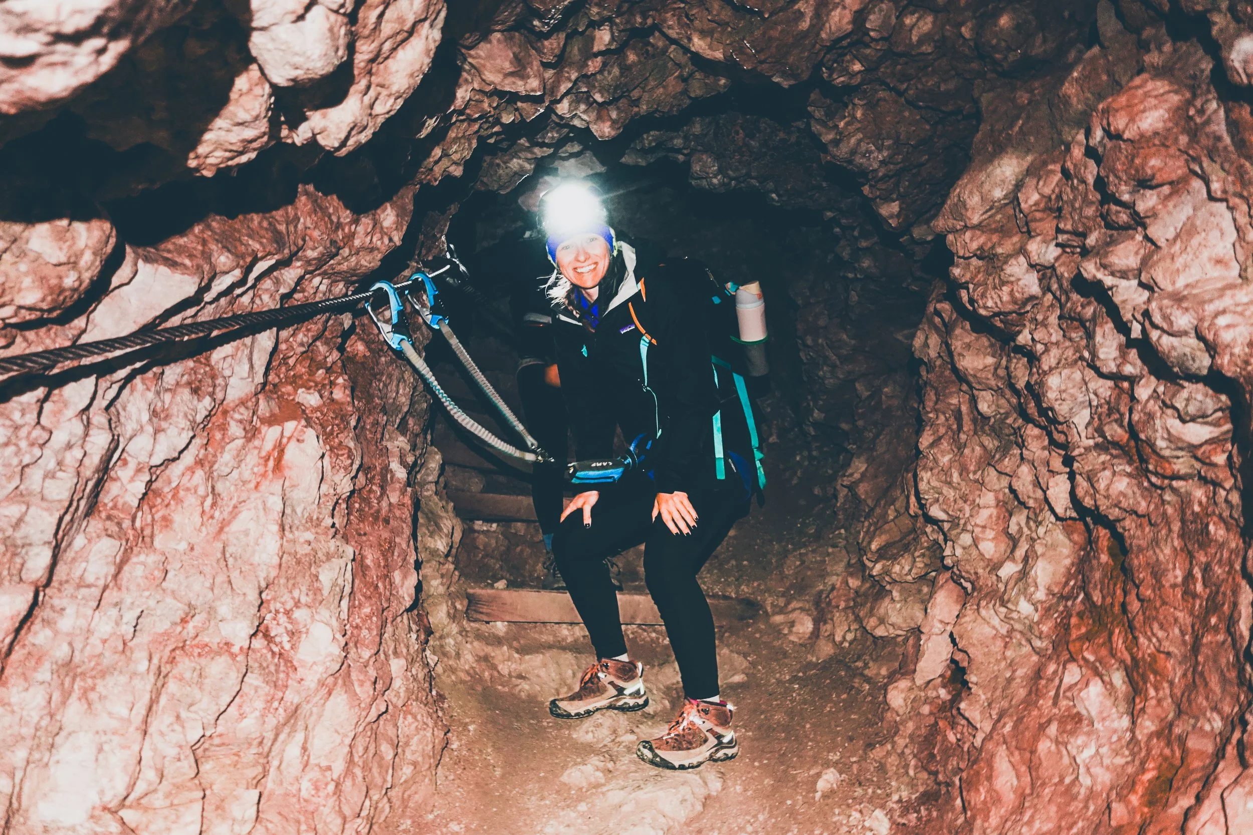 Woman in outdoor gear and headlamp smiling while crouching inside a narrow rocky cave or tunnel, holding onto a rope for support.