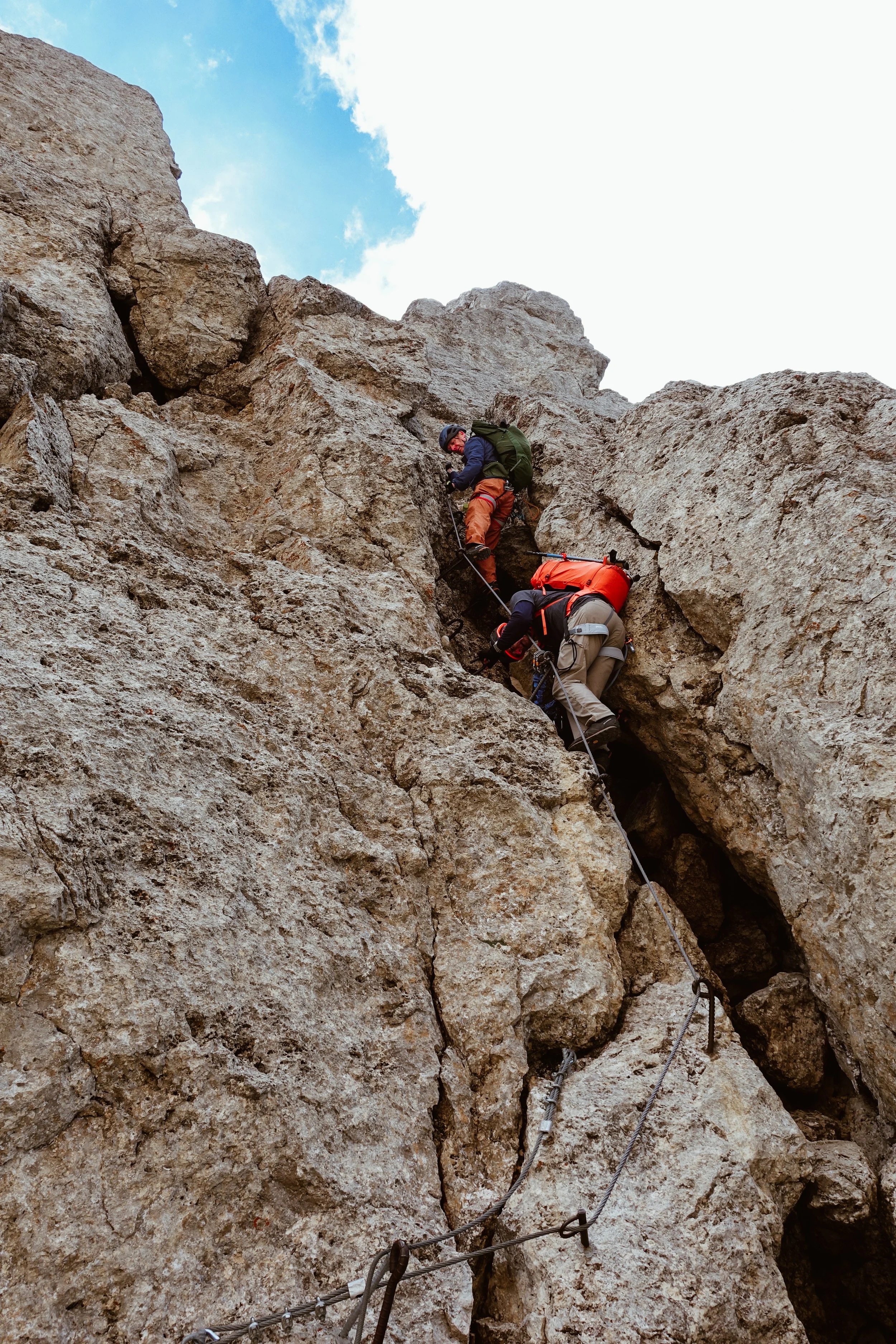 Two hikers climbing a rocky mountain face using a via ferrata protected by safety cables and helmets.