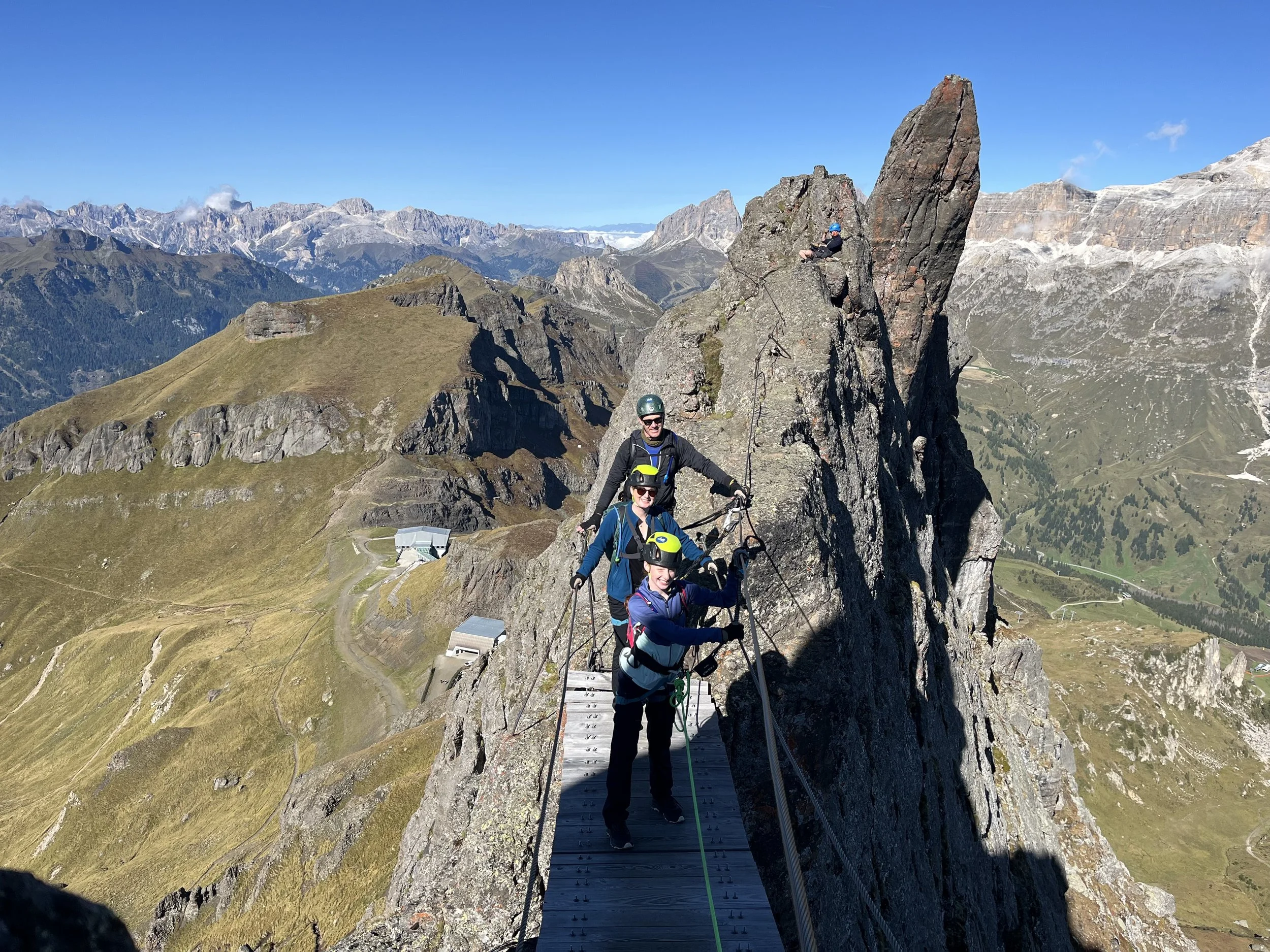 Four people wearing helmets and harnesses on a narrow mountain ridge with a scenic mountain landscape in the background. Via ferrata safari, trenches, Dolomites. 