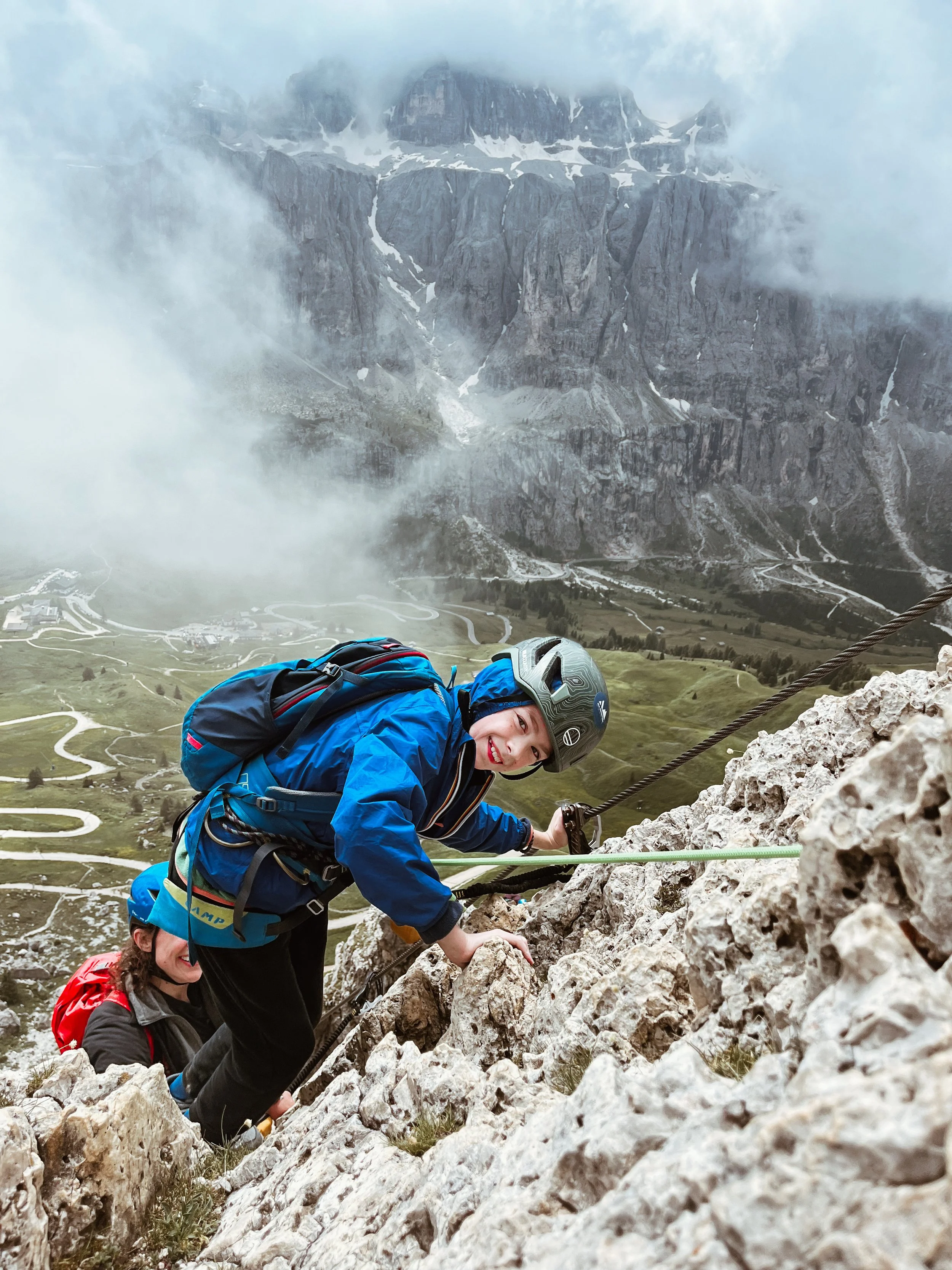 Two climbers, one woman and one man, ascending a rocky mountain face with safety ropes, against a backdrop of lush green valleys, winding roads, and dramatic mountains shrouded in clouds.