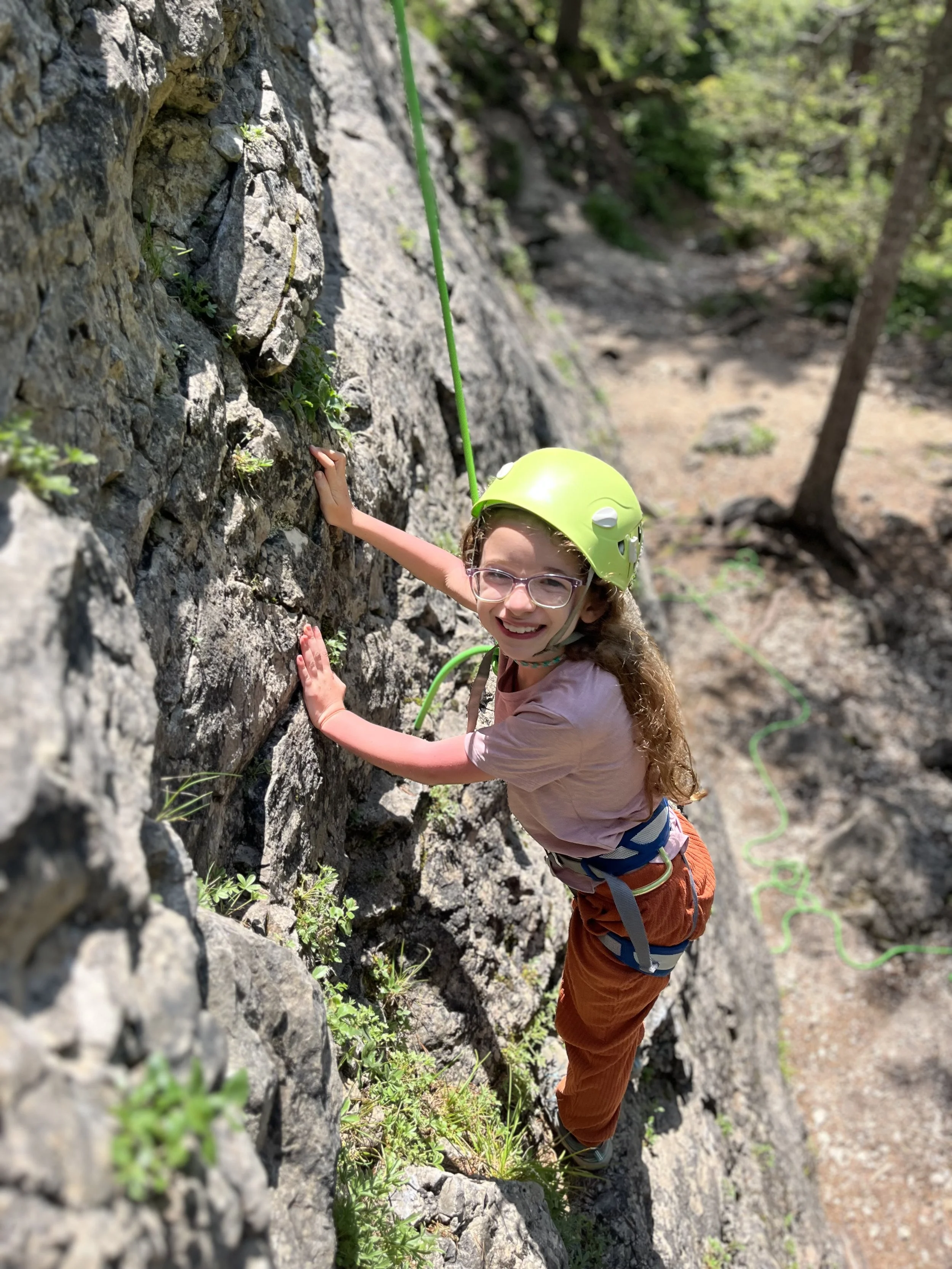 A young girl with glasses and curly hair rock climbing outdoors, wearing a yellow helmet and climbing harness, smiling at the camera.