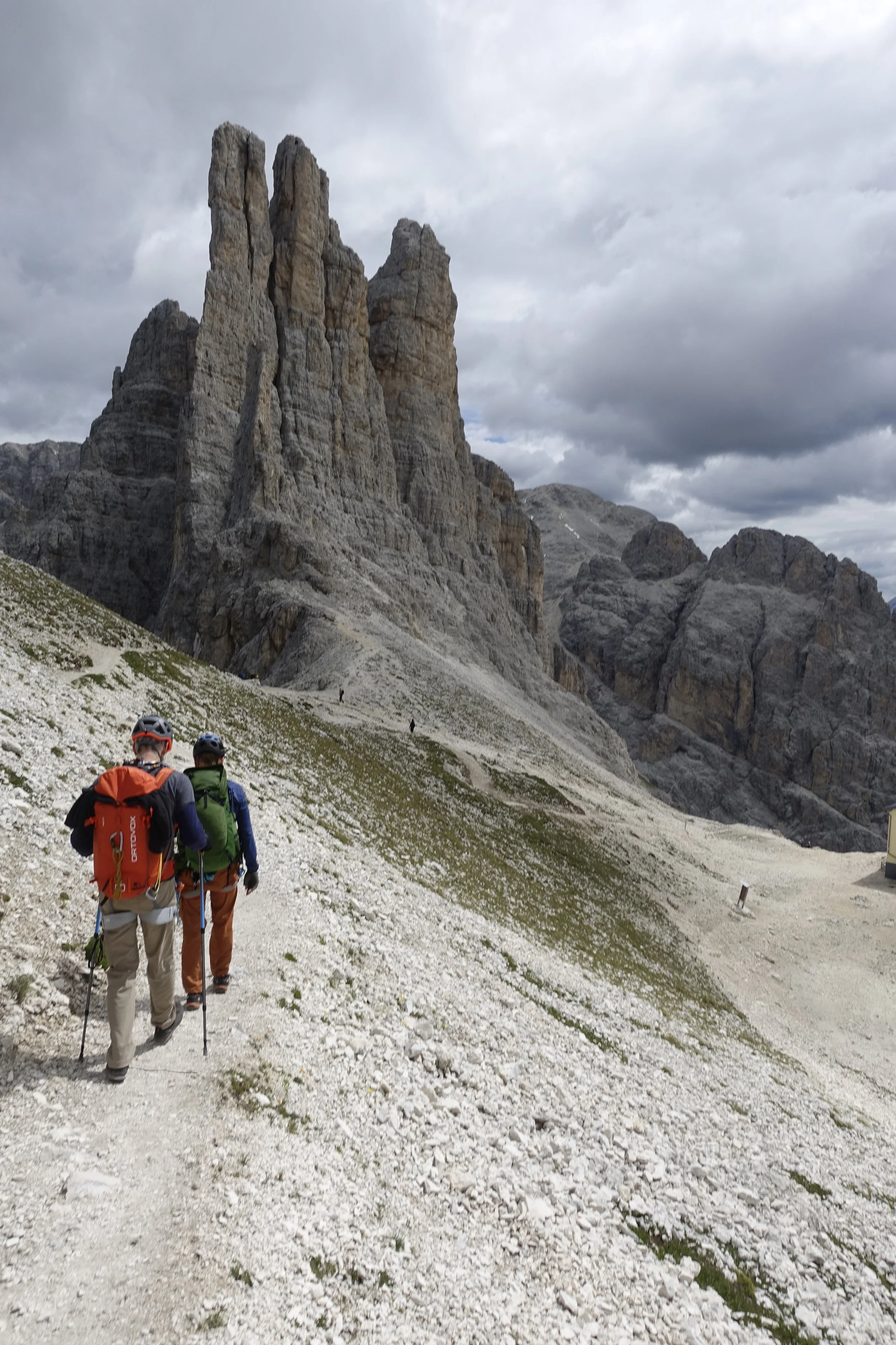 Two hikers wearing helmets and backpacks walking on a rocky mountain trail towards a large rock formation under cloudy skies, the three Vajolet Towers in the Gartl area. 