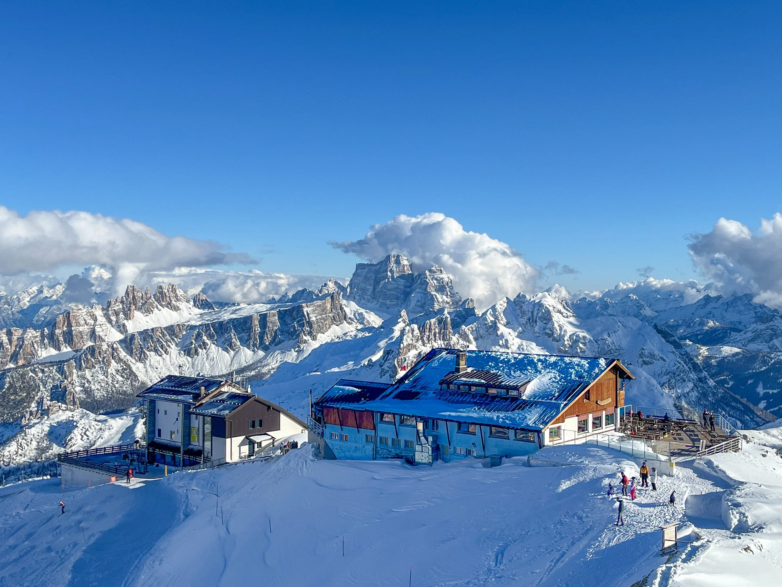 Snow-covered mountain landscape with a wooden lodge and a smaller building, with people skiing or snowboarding on the slope, under a clear blue sky.