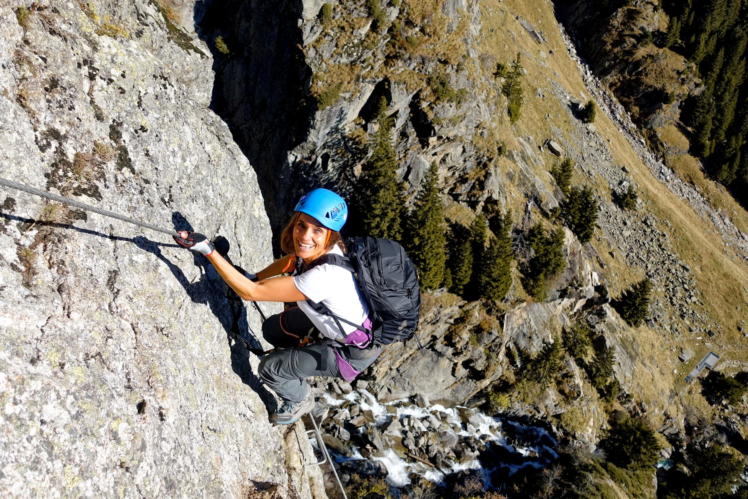 A woman wearing a blue helmet, white t-shirt, and climbing gear ascends a steep rock face, smiling at the camera with a backpack on her back, in a mountainous landscape with trees and a stream below.