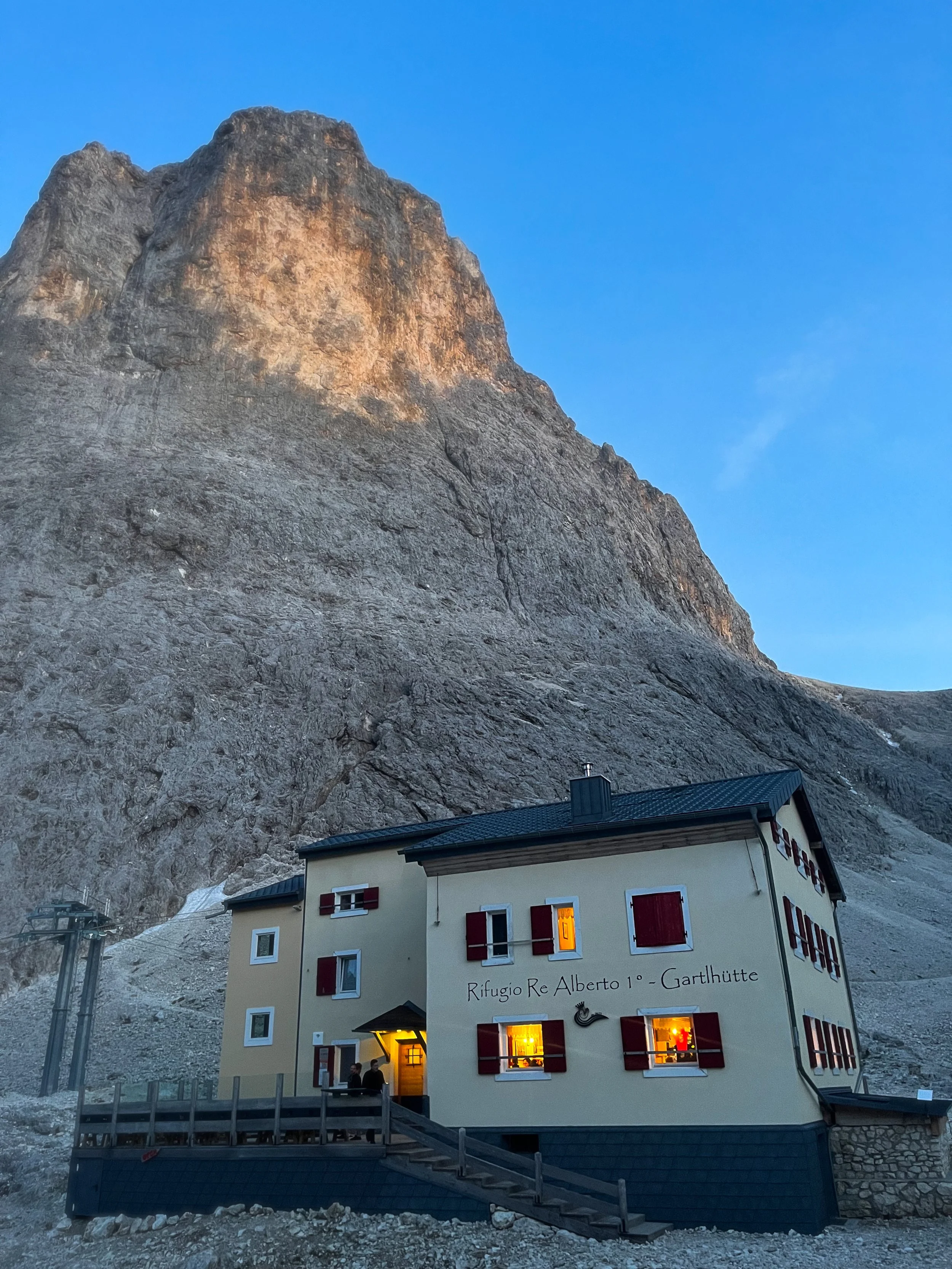 A mountain hut called Rifugio Re Alberto 1° Garthütte at the base of a large rocky mountain with the sky in the background. The hut has illuminated windows, red shutters, and a small porch with people standing near the entrance.