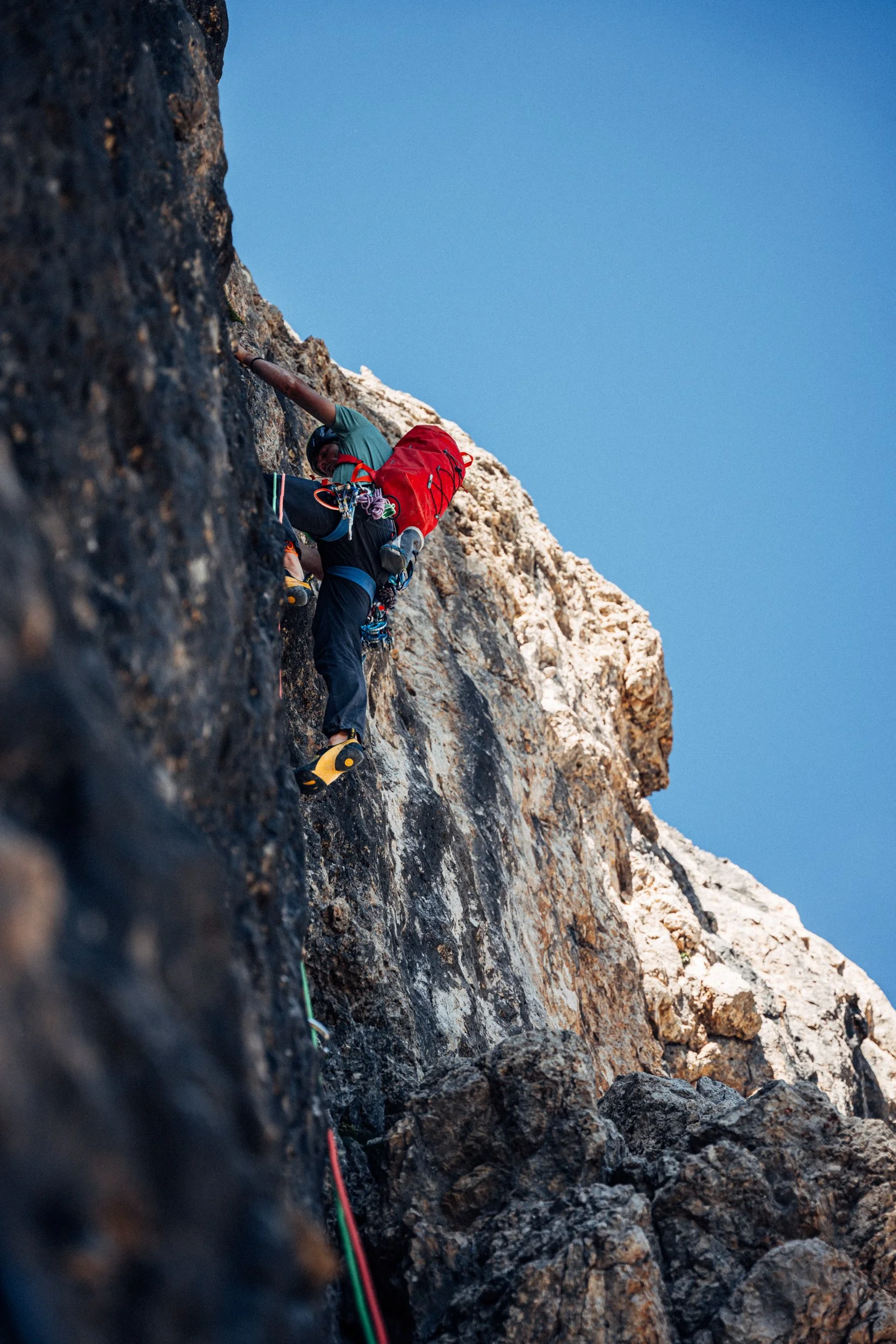 A person engaged in rock climbing on a steep cliff face, wearing climbing gear and using safety ropes, with a clear blue sky in the background.